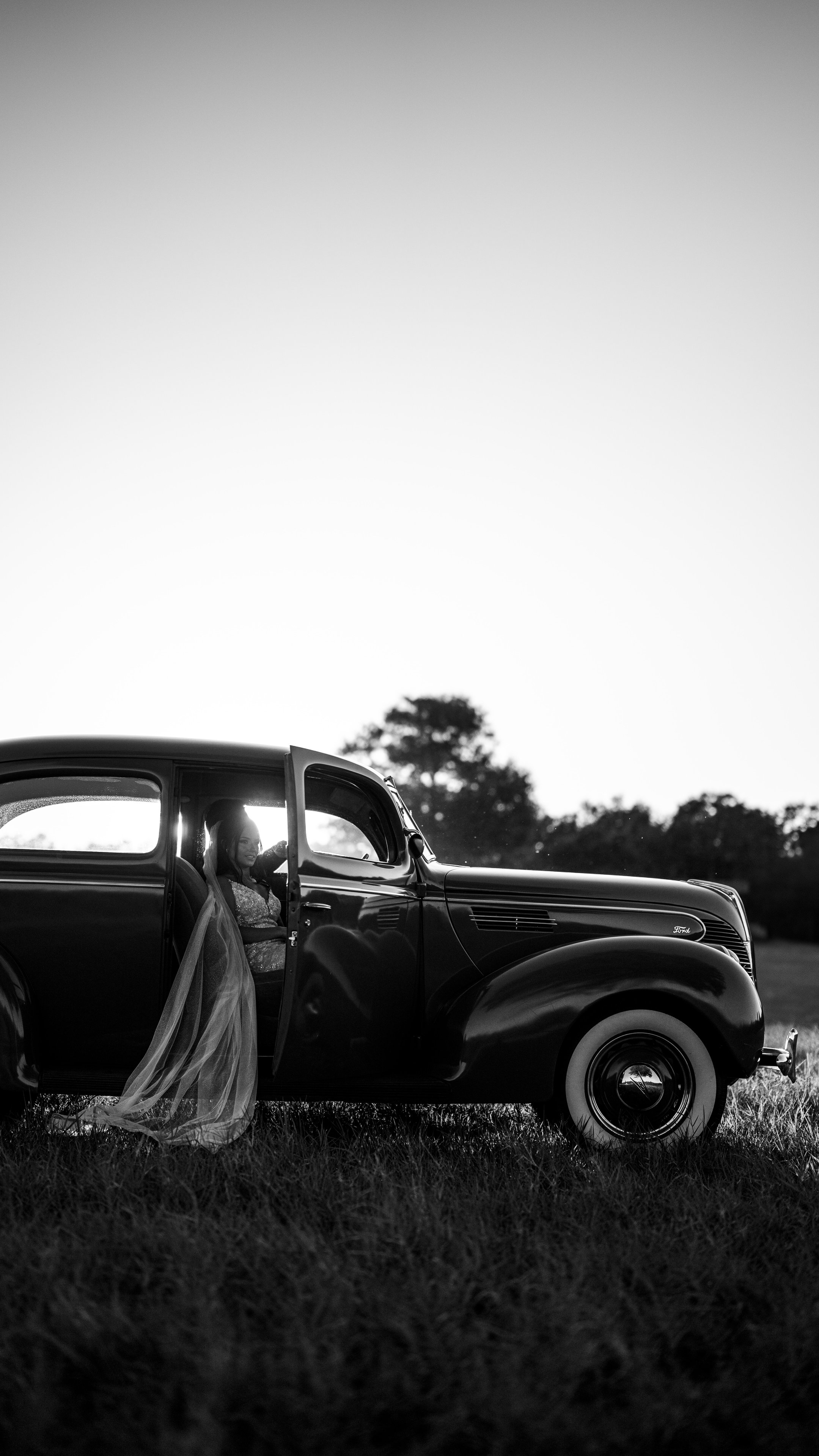 In black and white, a dark vintage car parked in a field with Kenadee's veil trailing beside it.