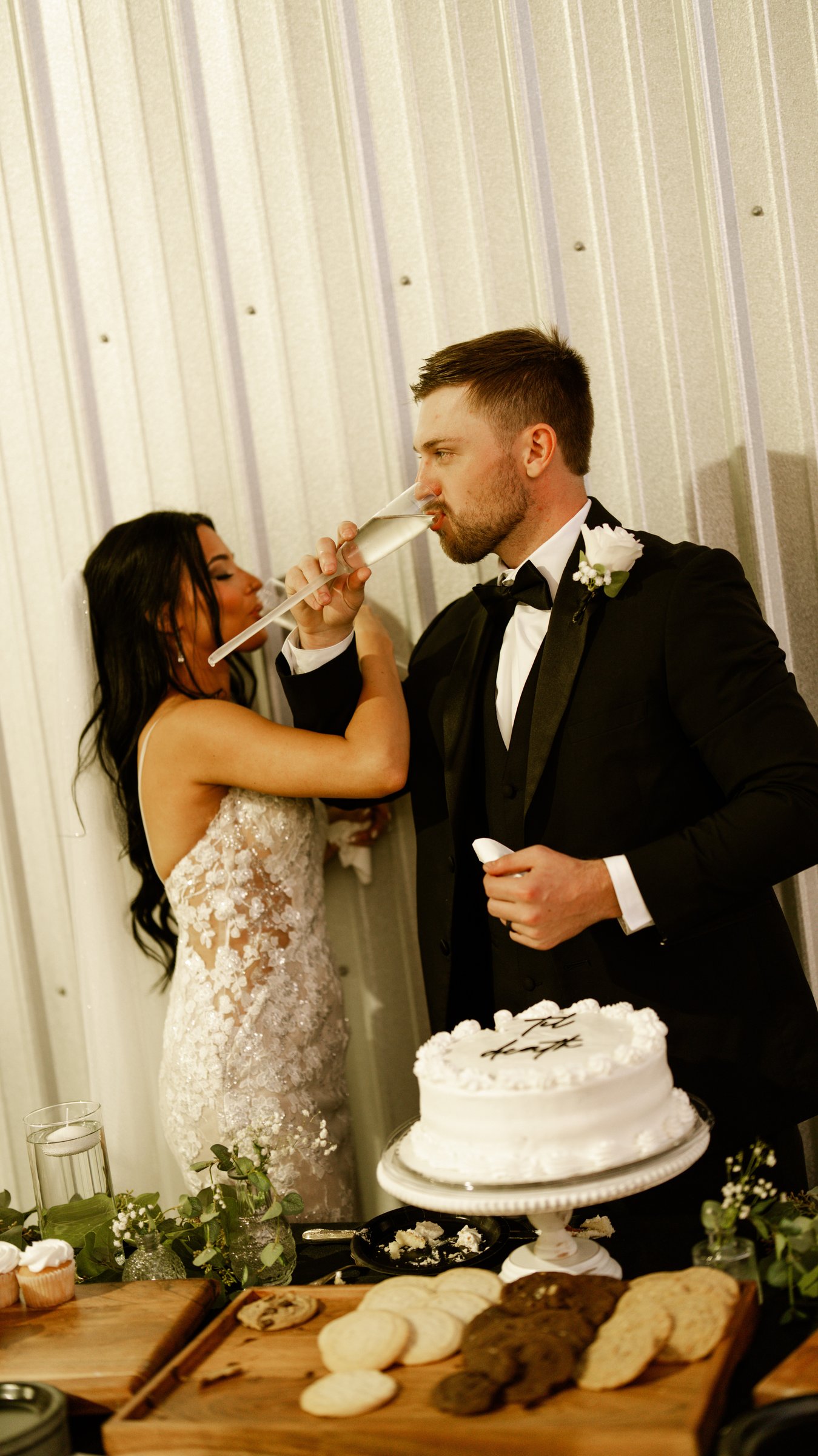 Kenadee and Corey drinking from glasses next to their white cake and a tray of cookies.