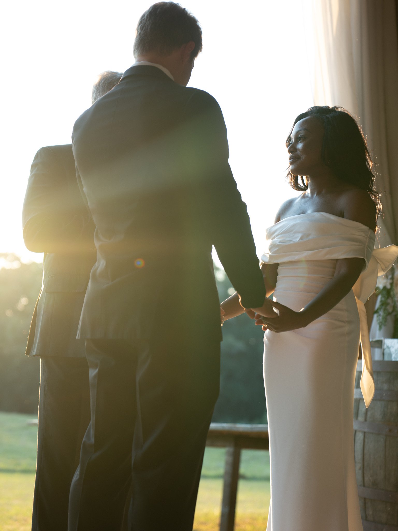 Jason, seen from behind, holds Ashley's hands while she looks off to the side in low sun, with another man standing just behind him.