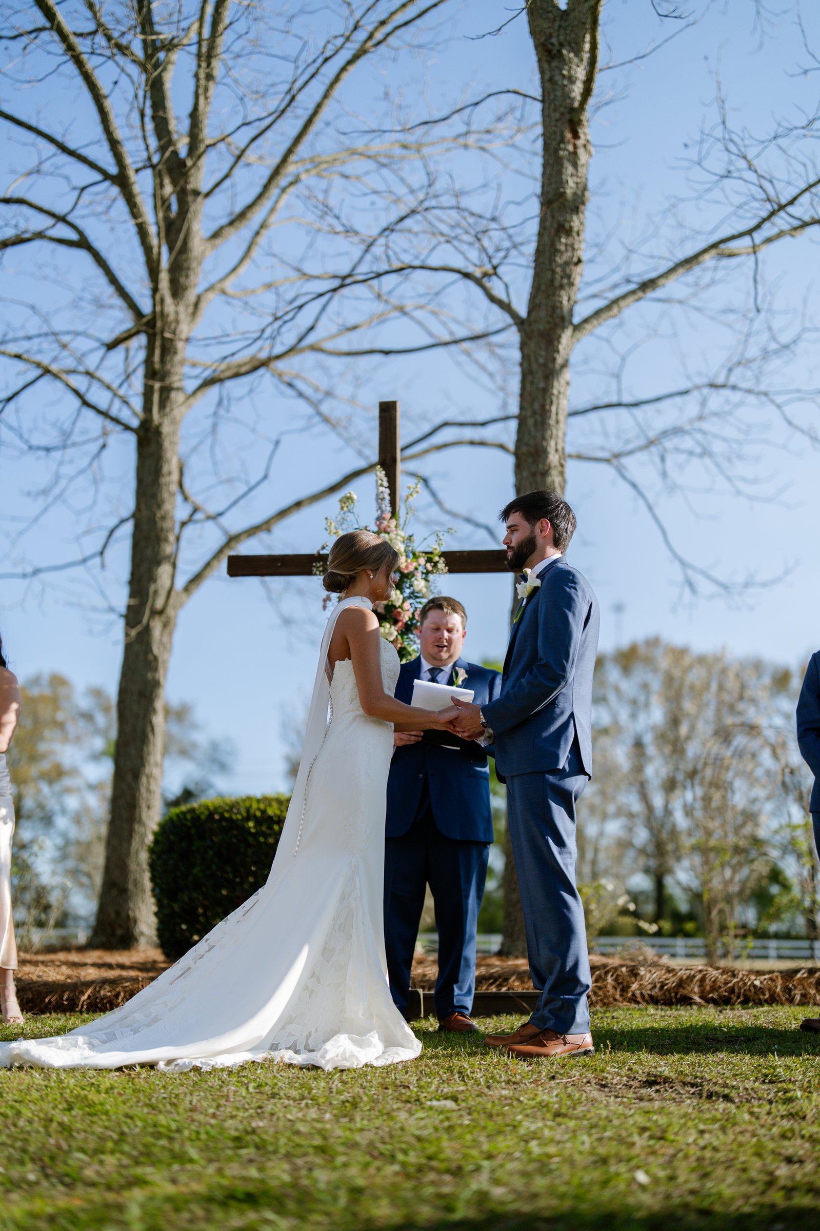 Joley and Matthew stand in front of a wooden cross facing a man in a dark suit, with bare trees and blue sky behind them.