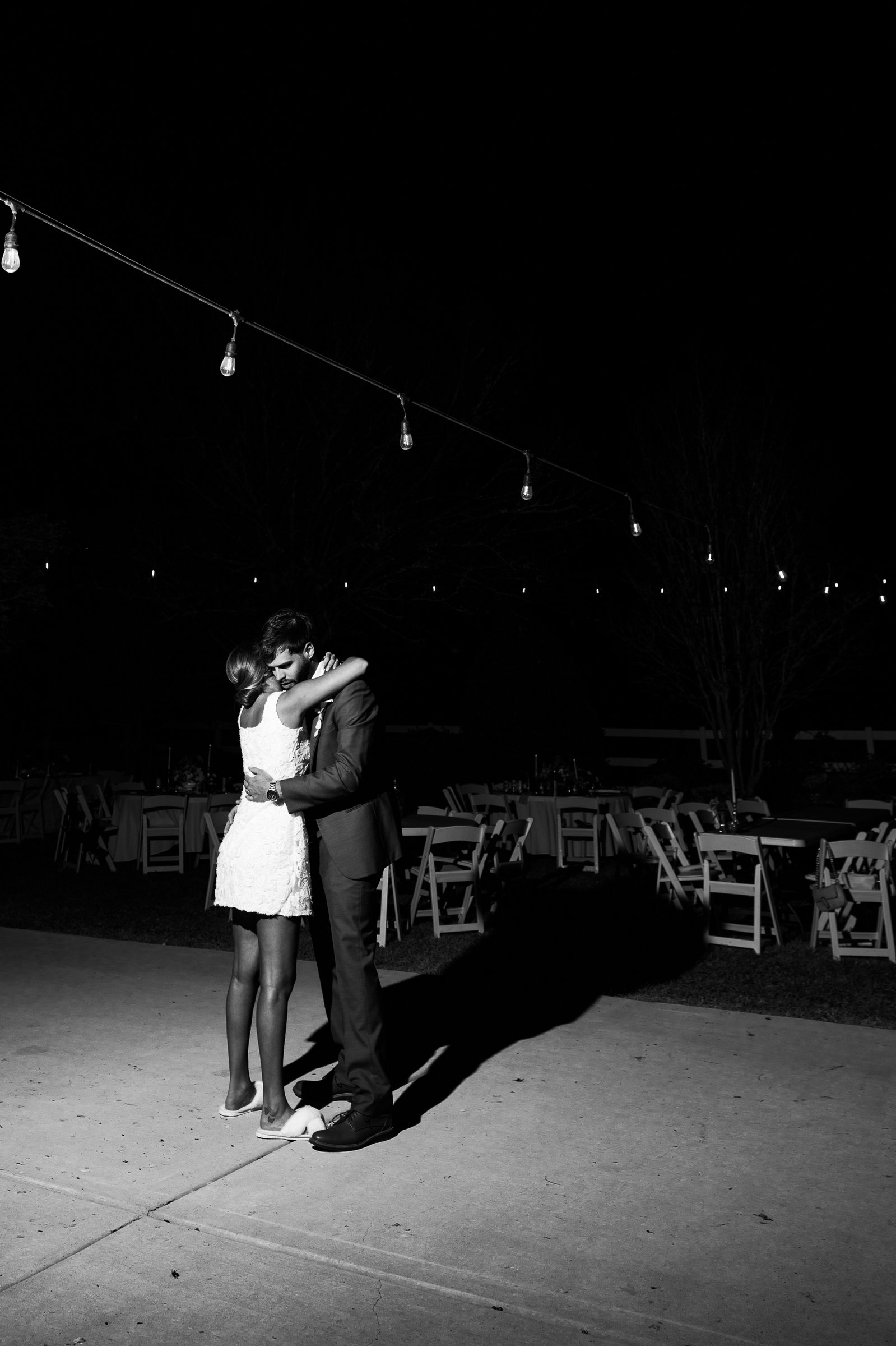 Black and white night photo of Joley and Matthew embracing on pavement under a string of lights.