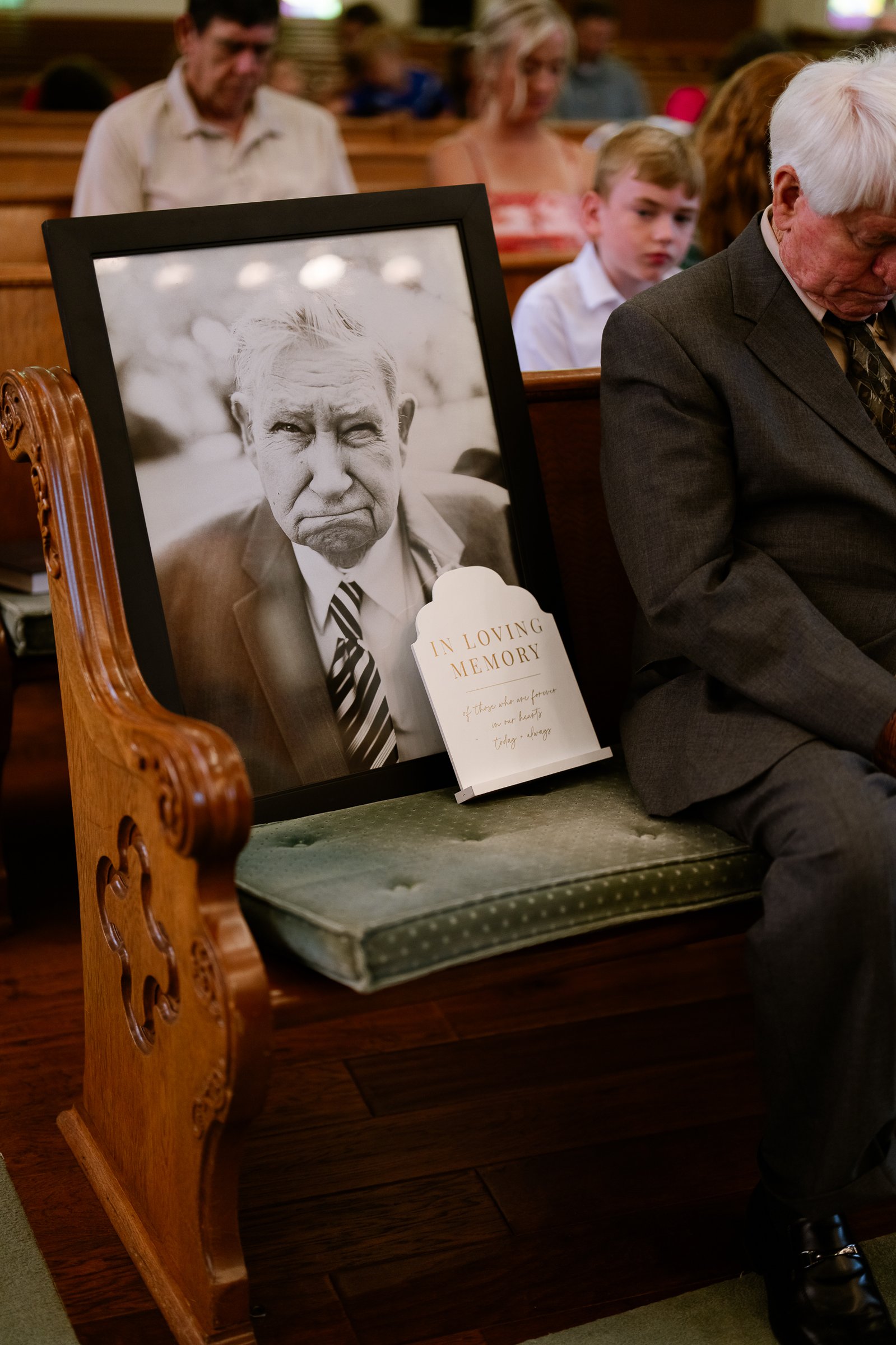 A framed black-and-white portrait of an older man with an In Loving Memory sign, set on a church pew beside a guest with his head bowed.