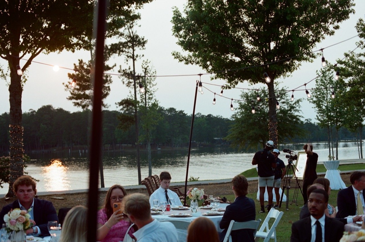 Reception tables beside the lake at New Water Farms, with the Midsummer crew and camera setup in the background, photographed on film.