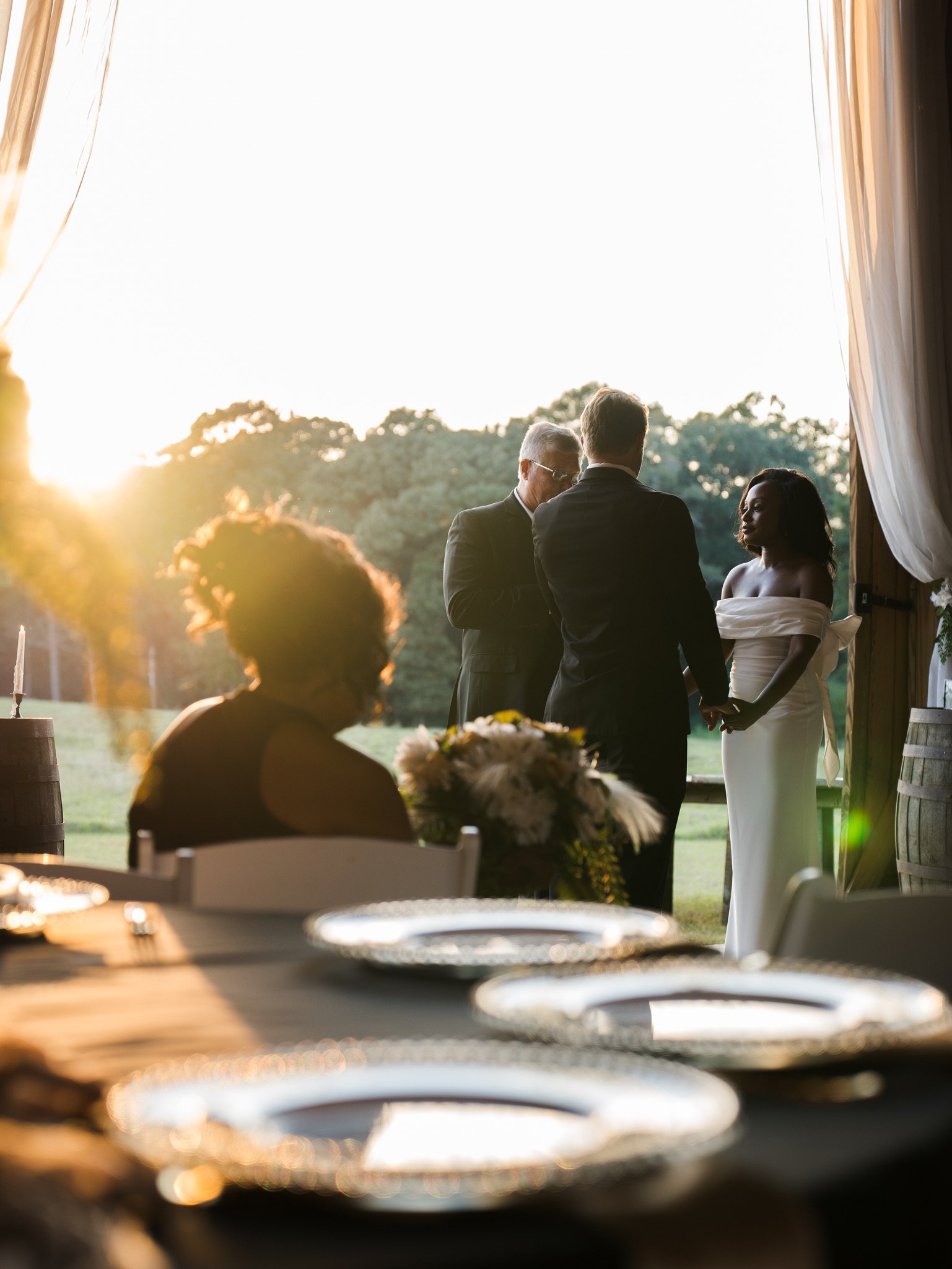 A guest seated at a table with silver plates in the foreground, while Ashley and Jason stand with a man at the edge of the barn in low sun.