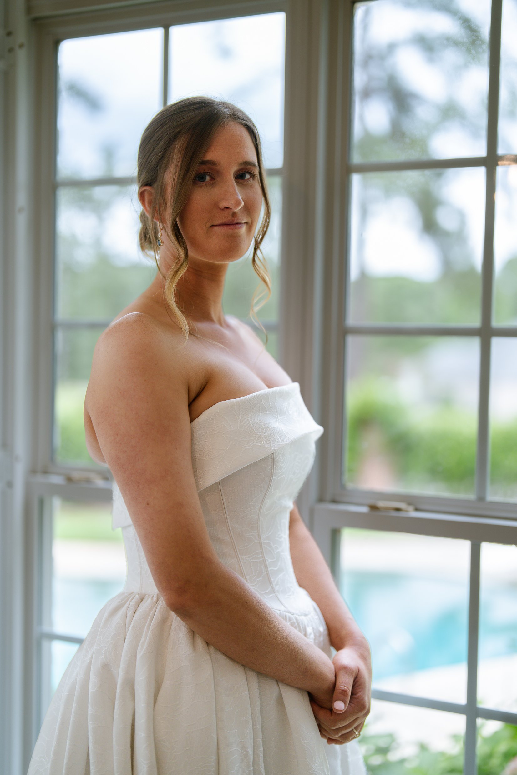 Elizabeth in her strapless gown looks toward the camera with her hands folded, between tall windows with trees and a pool outside.