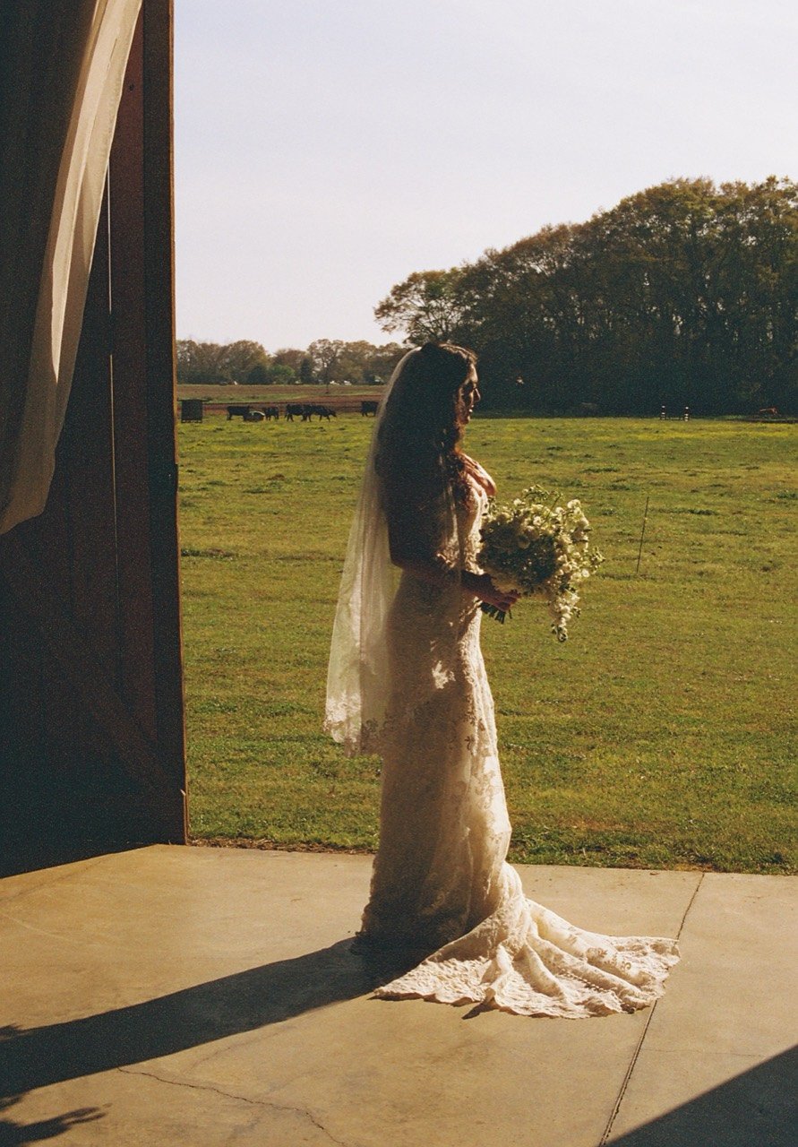 A full-length film portrait of Raley with her bouquet, lace train and veil in the sunlit doorway at Dalrymple Farms.
