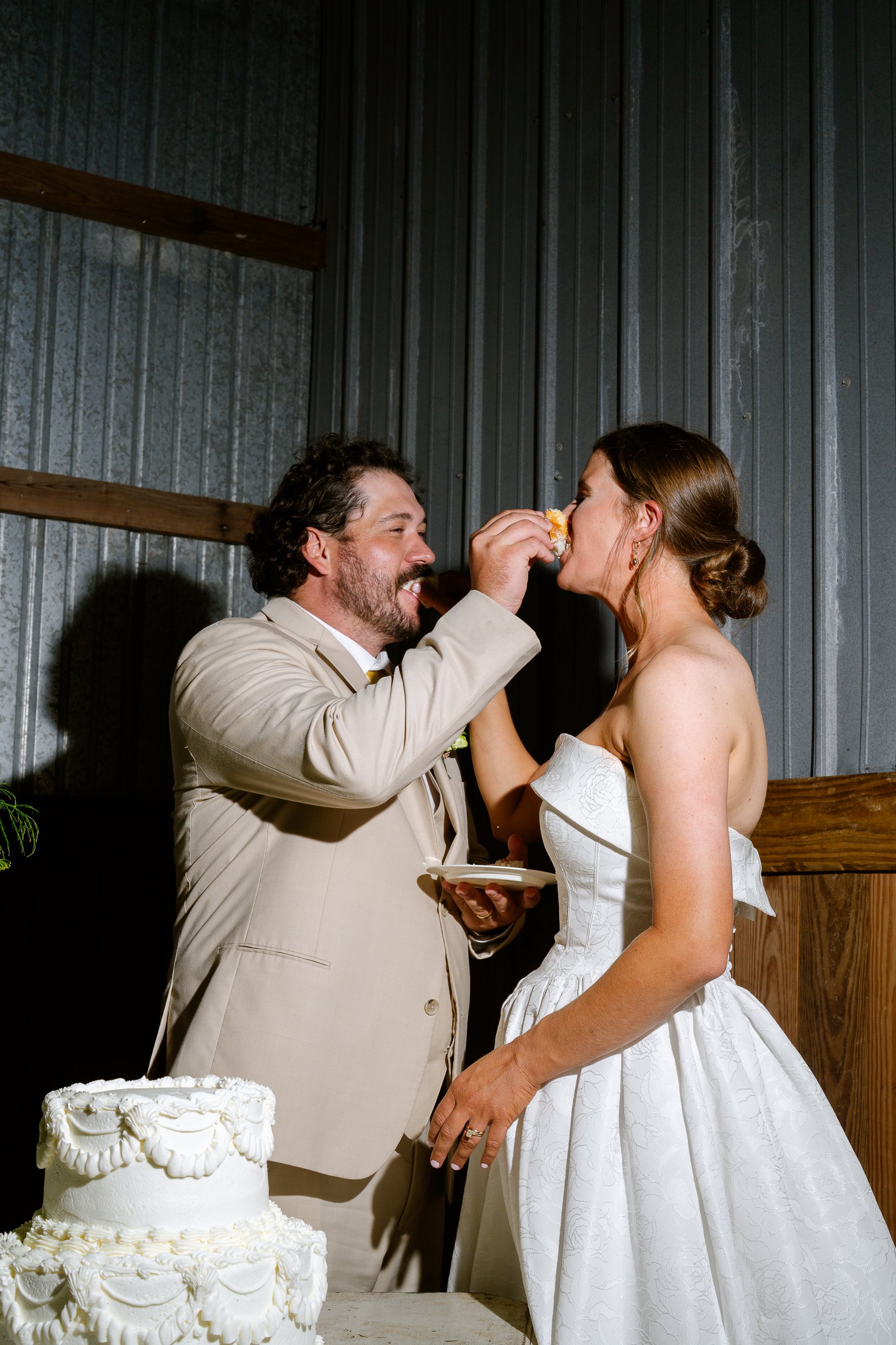 Dustin lifts a piece of cake to Elizabeth's mouth beside the cake table in the barn.