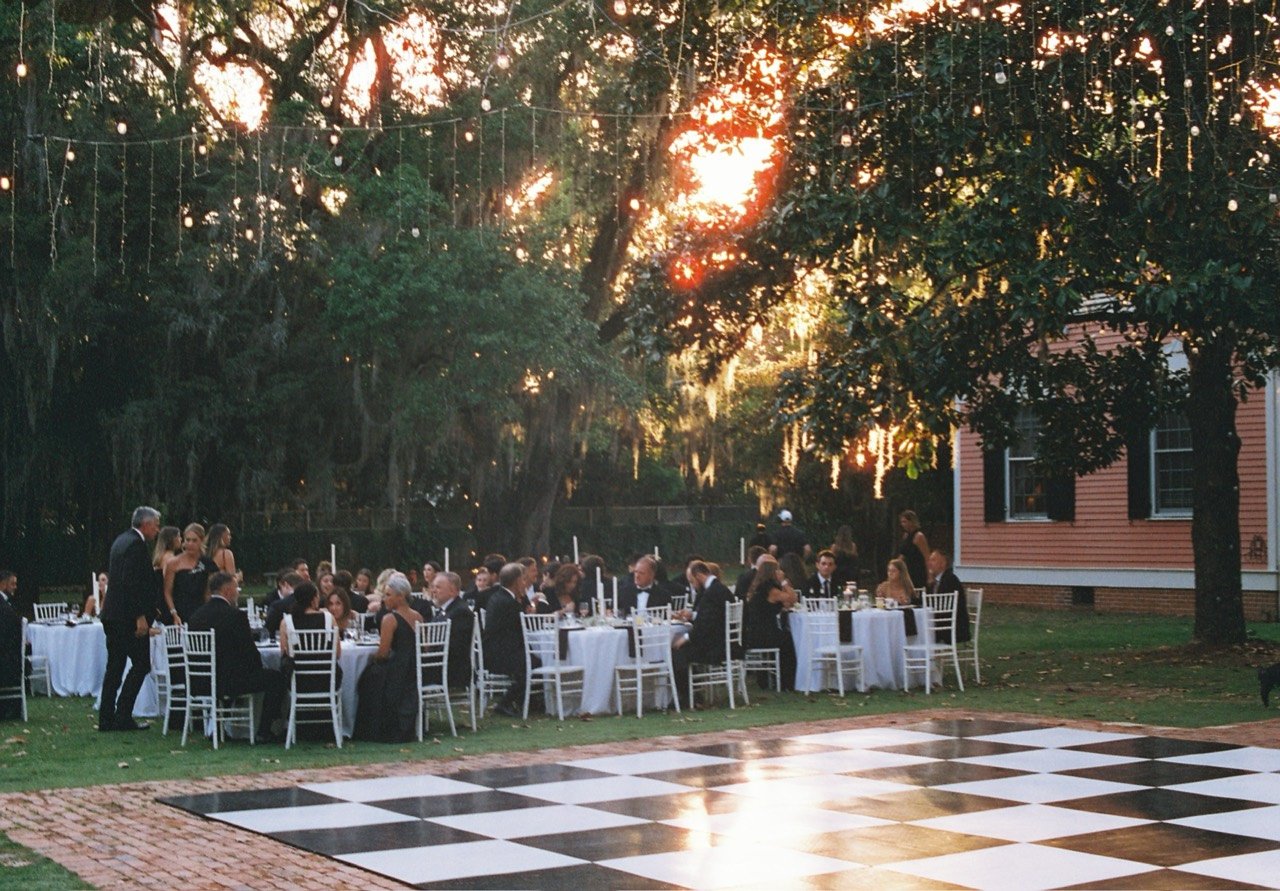 Guests at outdoor dinner tables beside a checkerboard dance floor beneath oak trees and string lights at Emily and Timothy’s wedding, photographed on film.