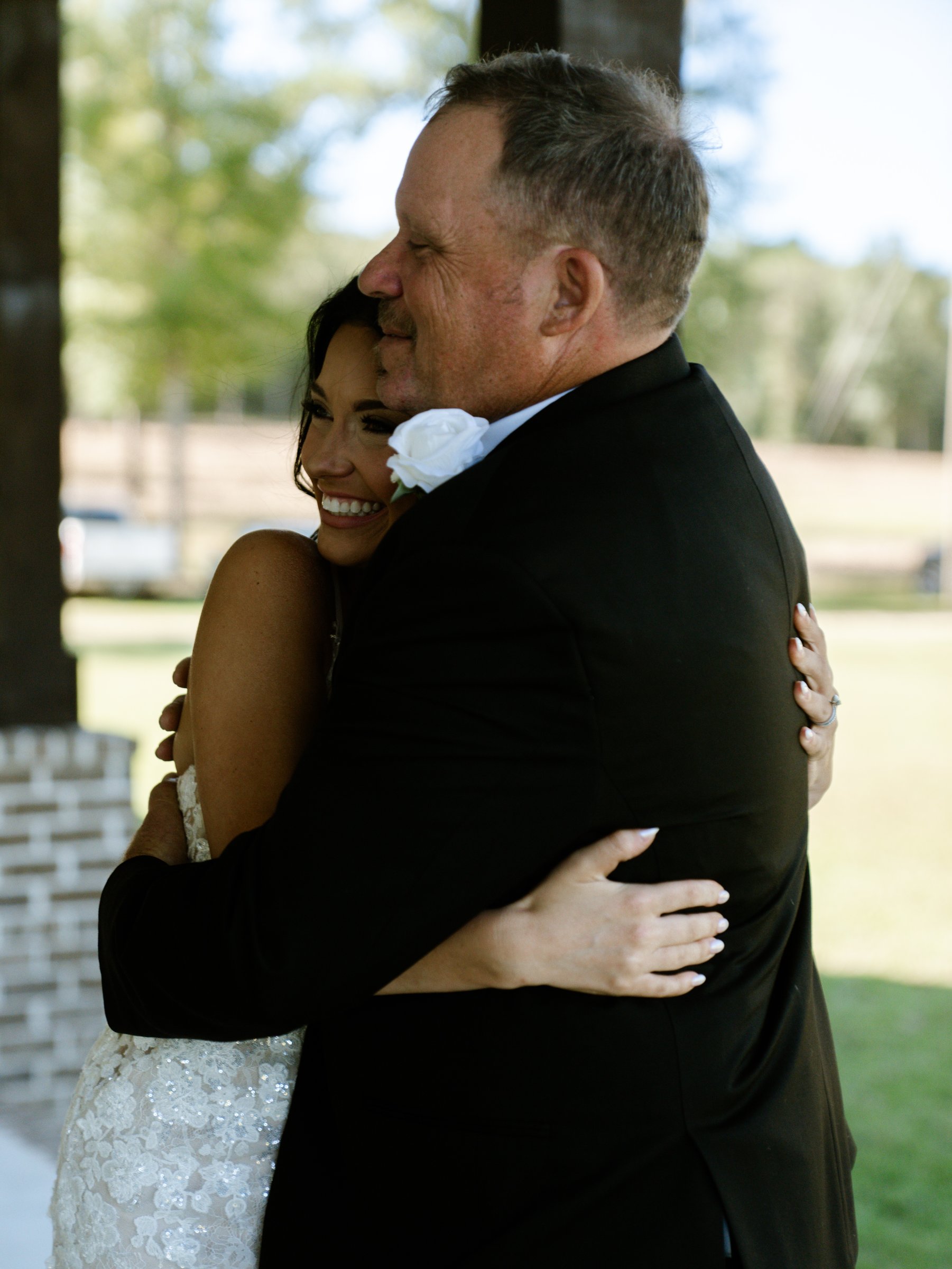 Kenadee in her dress hugging a man in a black tuxedo with a white flower on his lapel, outdoors.