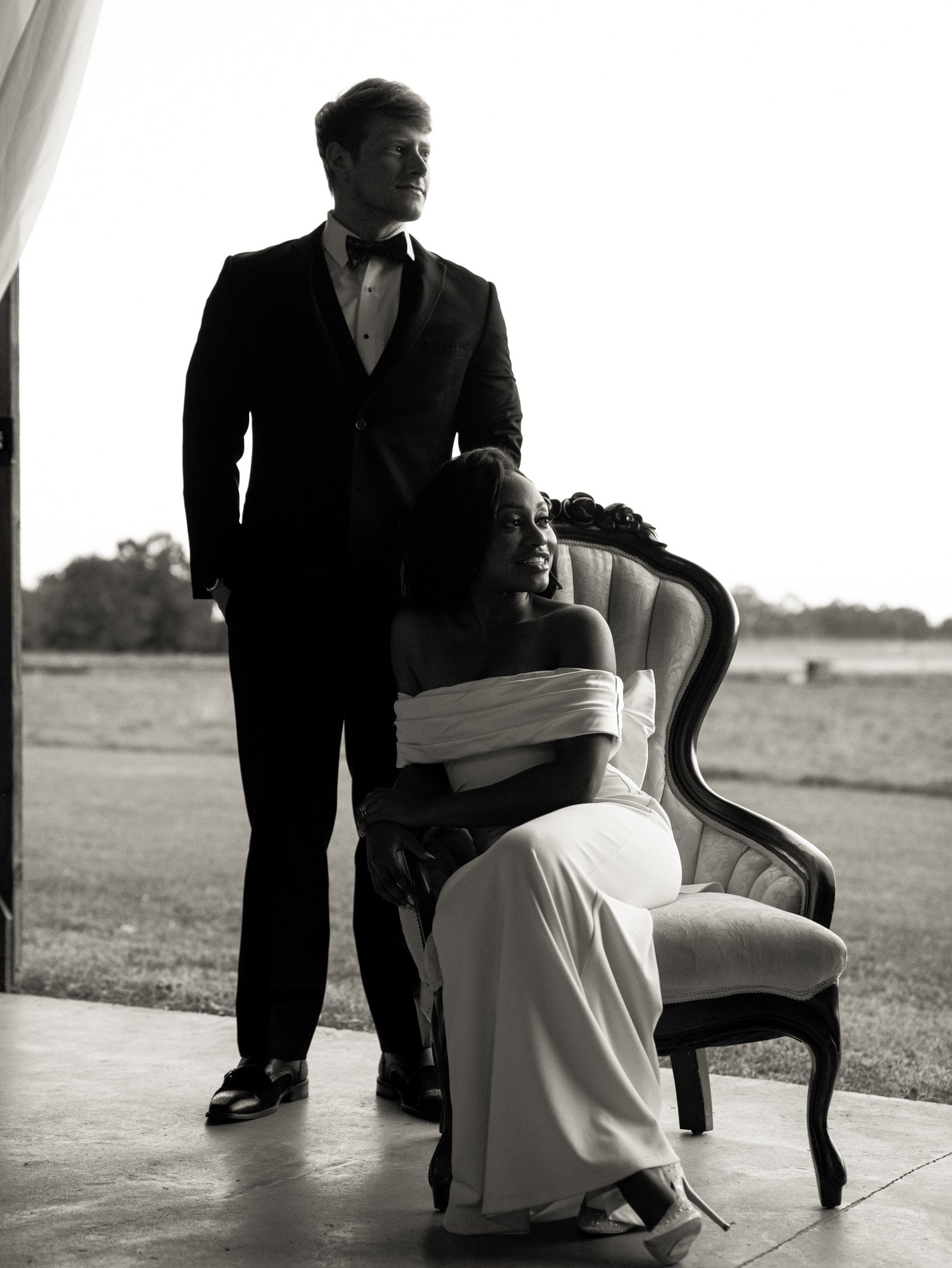 Jason stands behind Ashley, who sits in an upholstered chair, with a field behind them and a bright sky, in black and white.