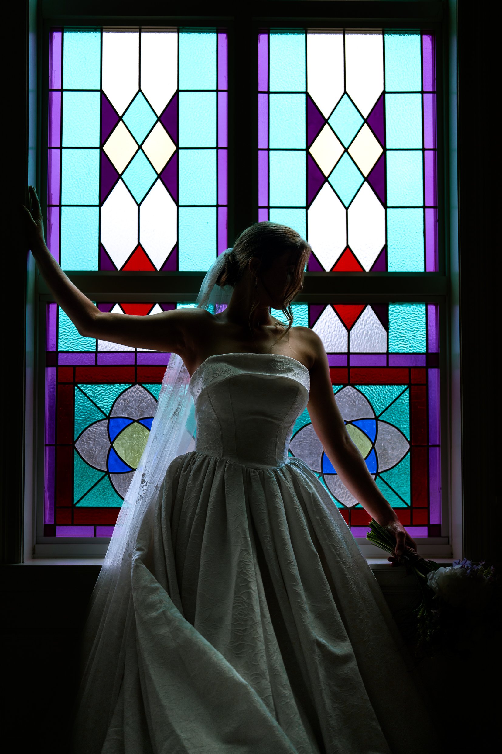 Elizabeth in silhouette with one hand on the window frame, her gown glowing against purple, blue and red stained glass.