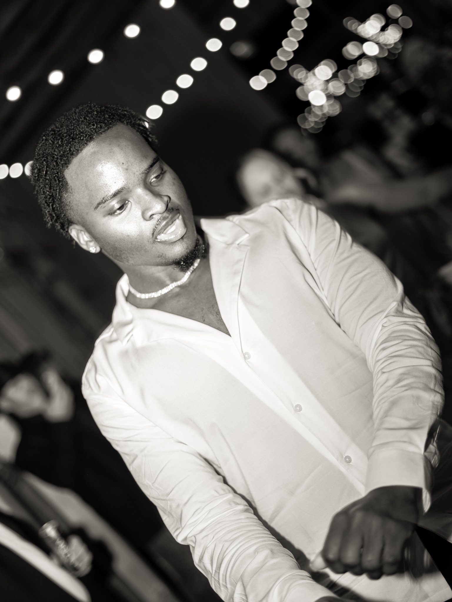 In black and white, a man in an open white shirt and a pearl necklace looks down, with string lights blurred behind him.