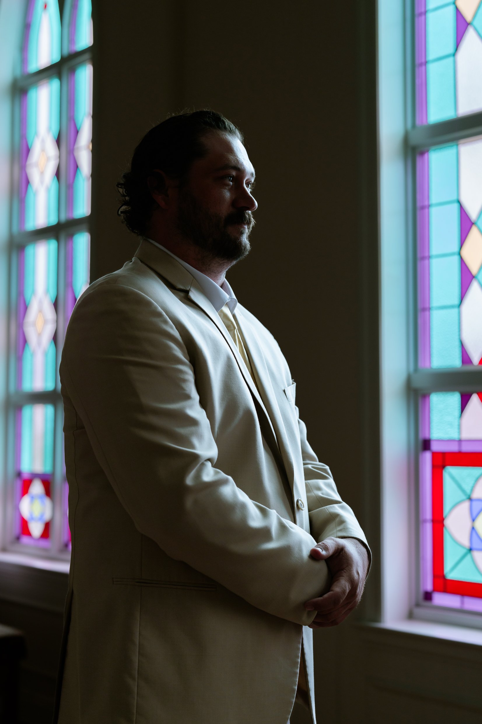 Dustin stands in profile beside a stained glass window with his hands folded, lit from the side.
