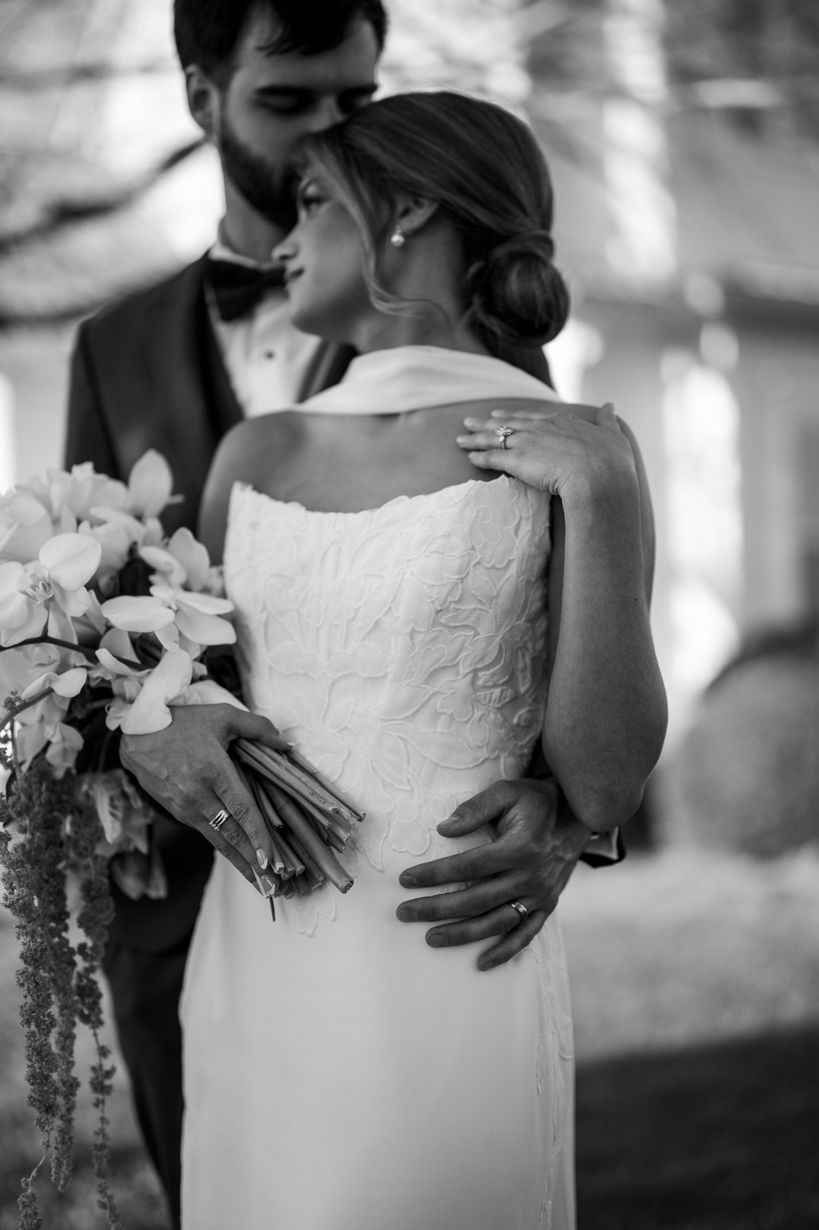 Black and white close view of Joley and Matthew embracing, her hands holding a bouquet in front of her and the background soft.