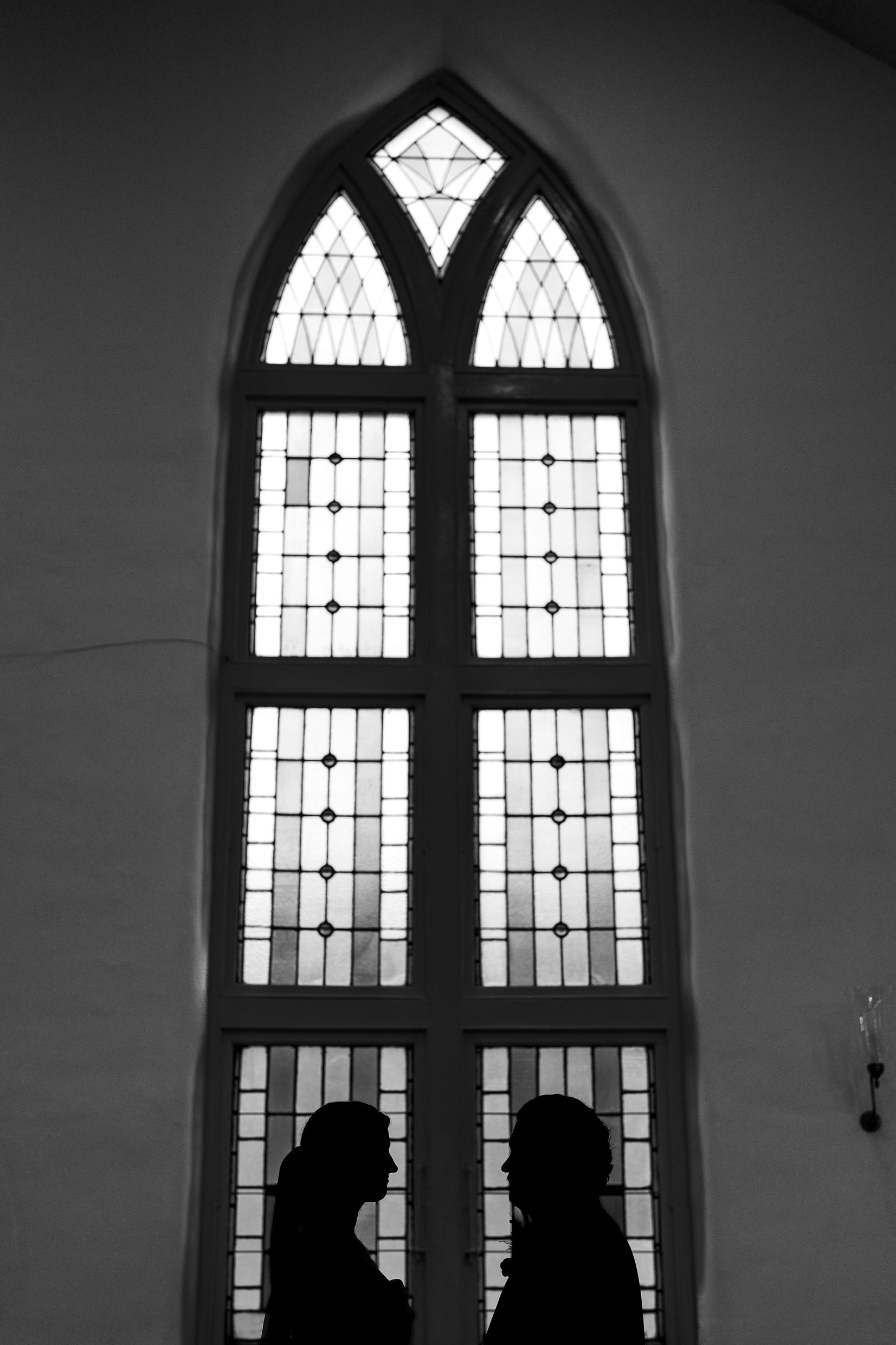 Elizabeth and Dustin stand as dark silhouettes at the bottom of a tall arched church window, in black and white.