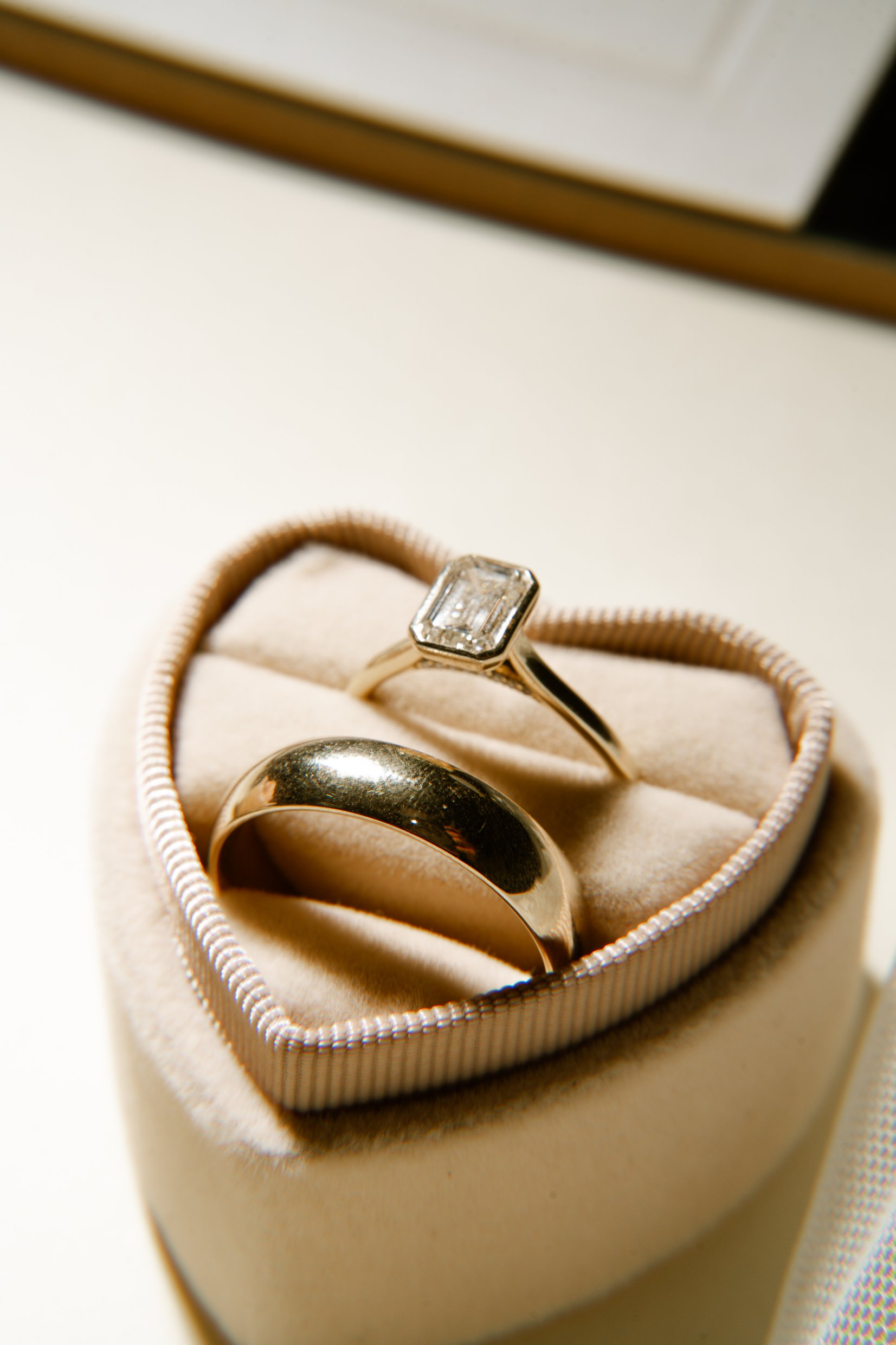 An emerald-cut diamond ring and a gold band side by side in a heart-shaped ring box.