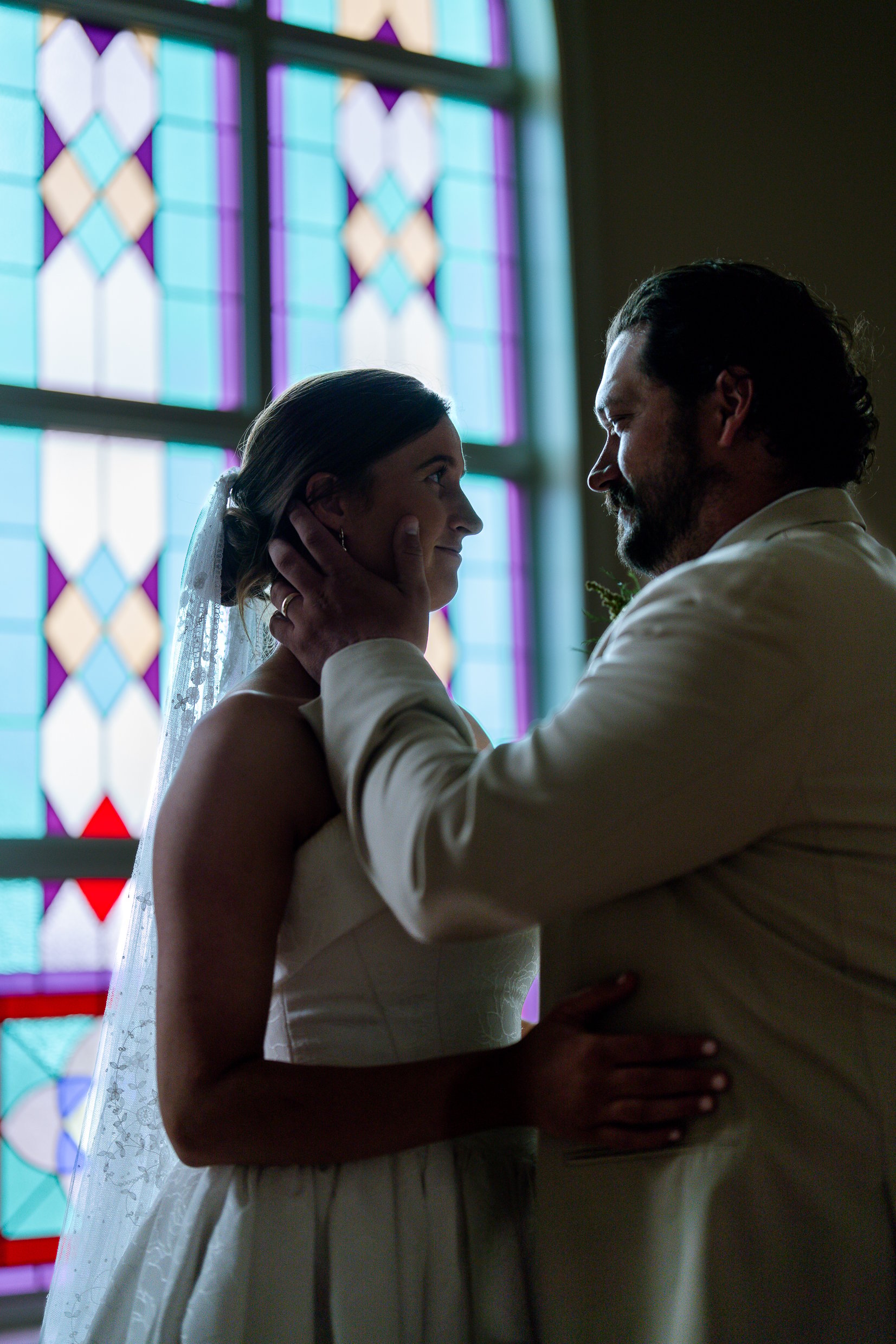 Elizabeth and Dustin stand close in front of a stained glass window, his hand on her cheek as they look at each other.