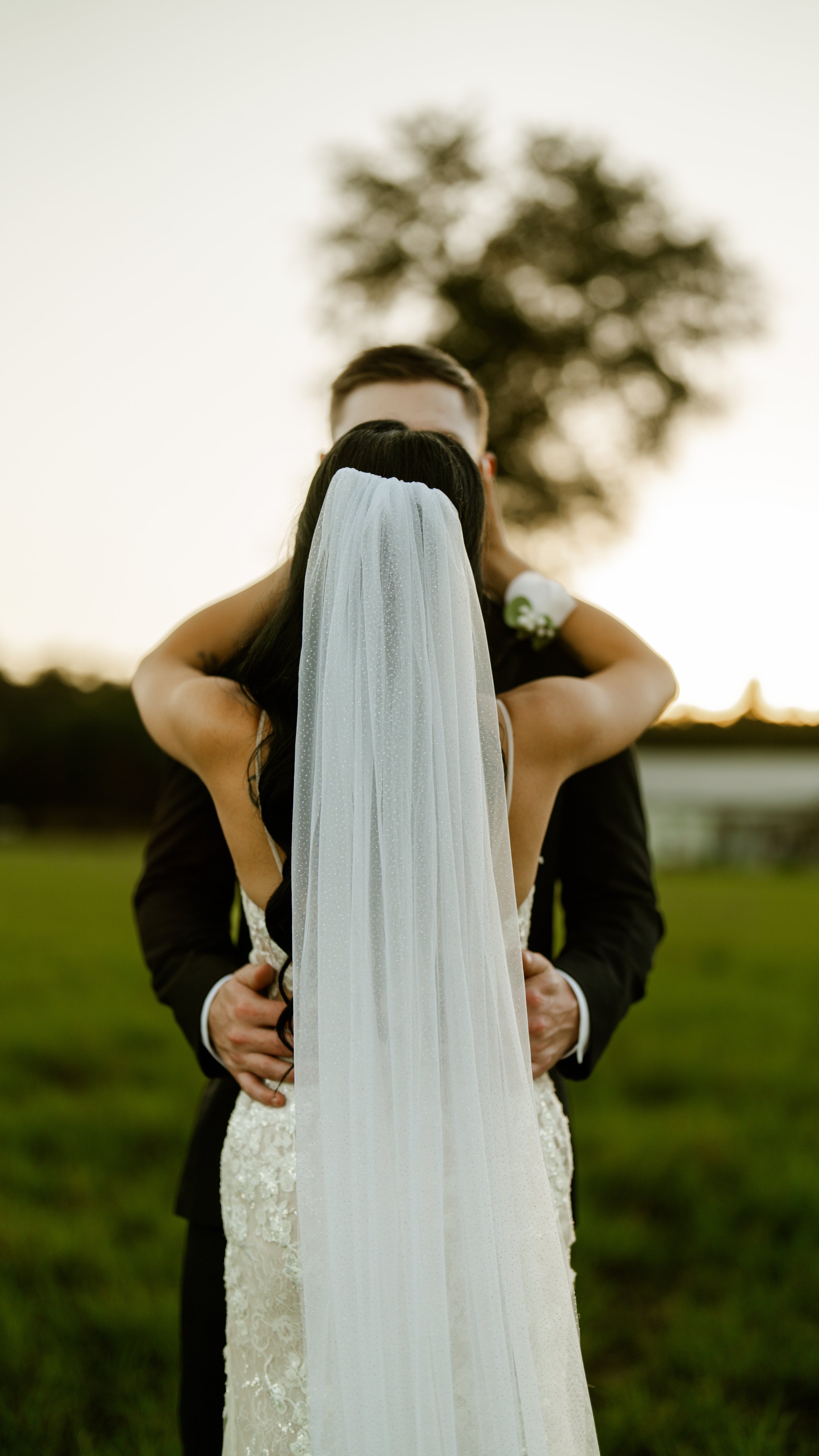 Kenadee with her back to the camera in her veil, with Corey's arms around her and a tree out of focus behind them.
