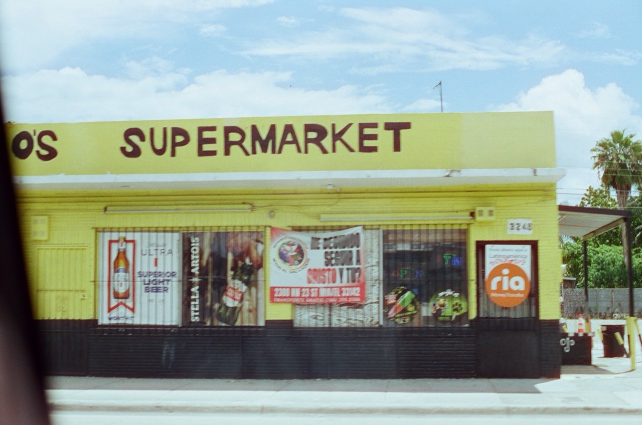A yellow supermarket storefront seen through a vehicle window during Raley’s Miami trip, photographed on film.