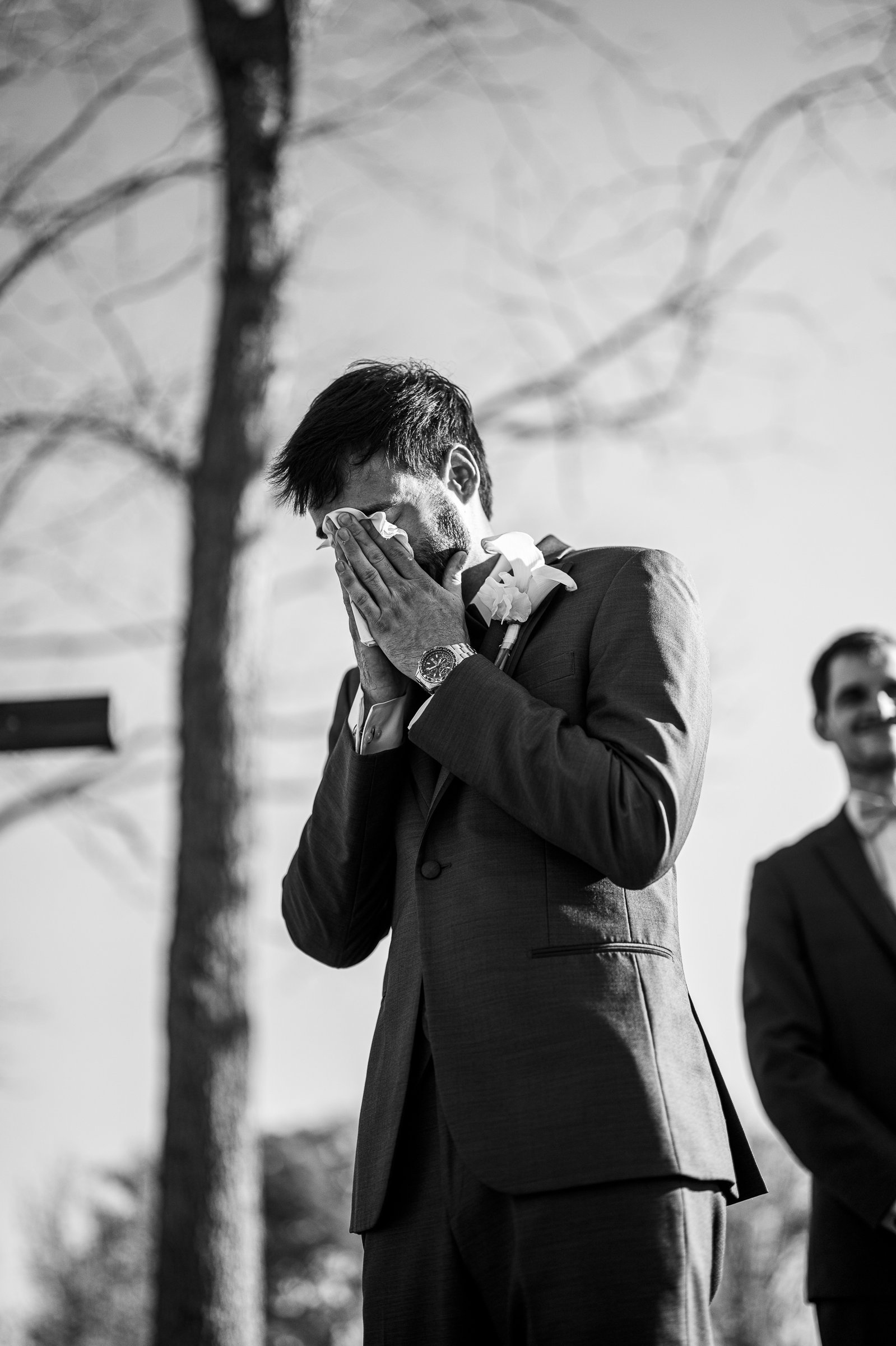 Black and white photo of a man in a suit covering his face with both hands, with a cross and another man behind him.