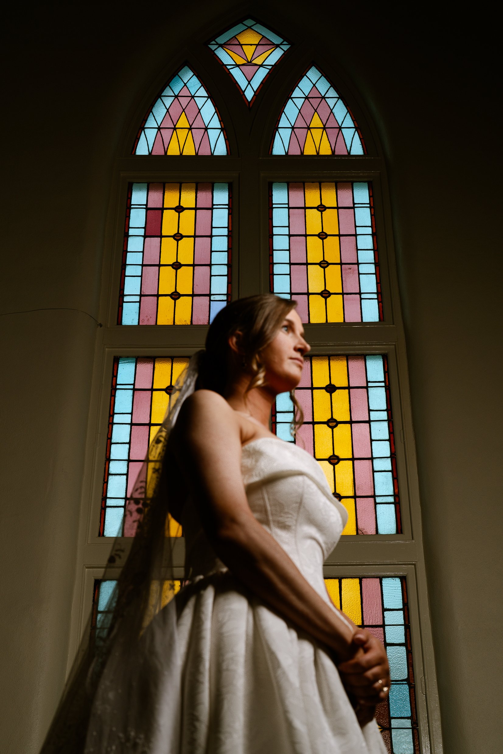 Elizabeth, lit from the side with her hands clasped, looks up in front of the church's tall stained glass window.