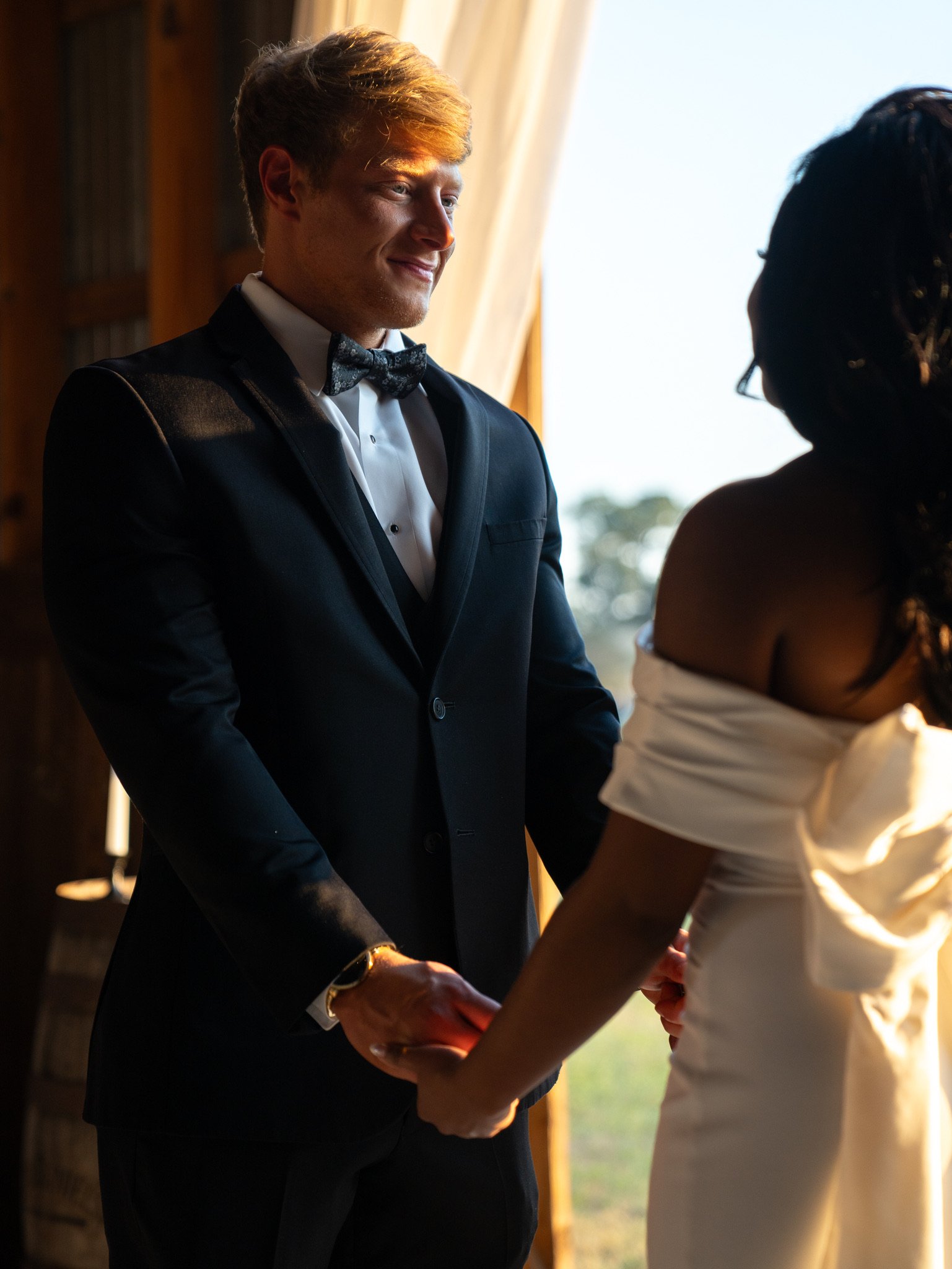 Jason smiles as he holds Ashley's hand in the barn opening, with a bright sky behind him.