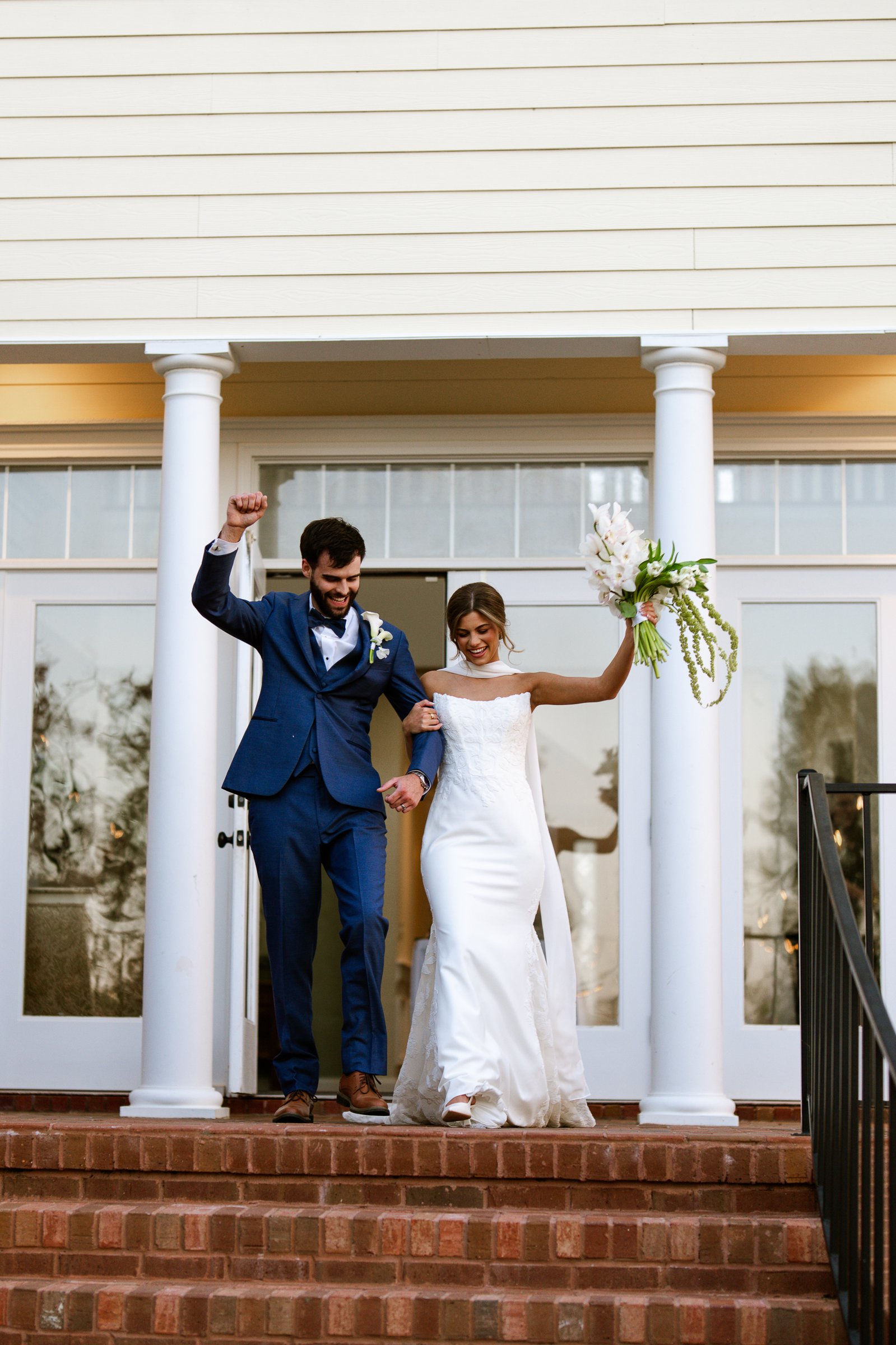 Joley and Matthew step down brick stairs from a white-columned porch, Joley raising her bouquet and Matthew raising a fist.