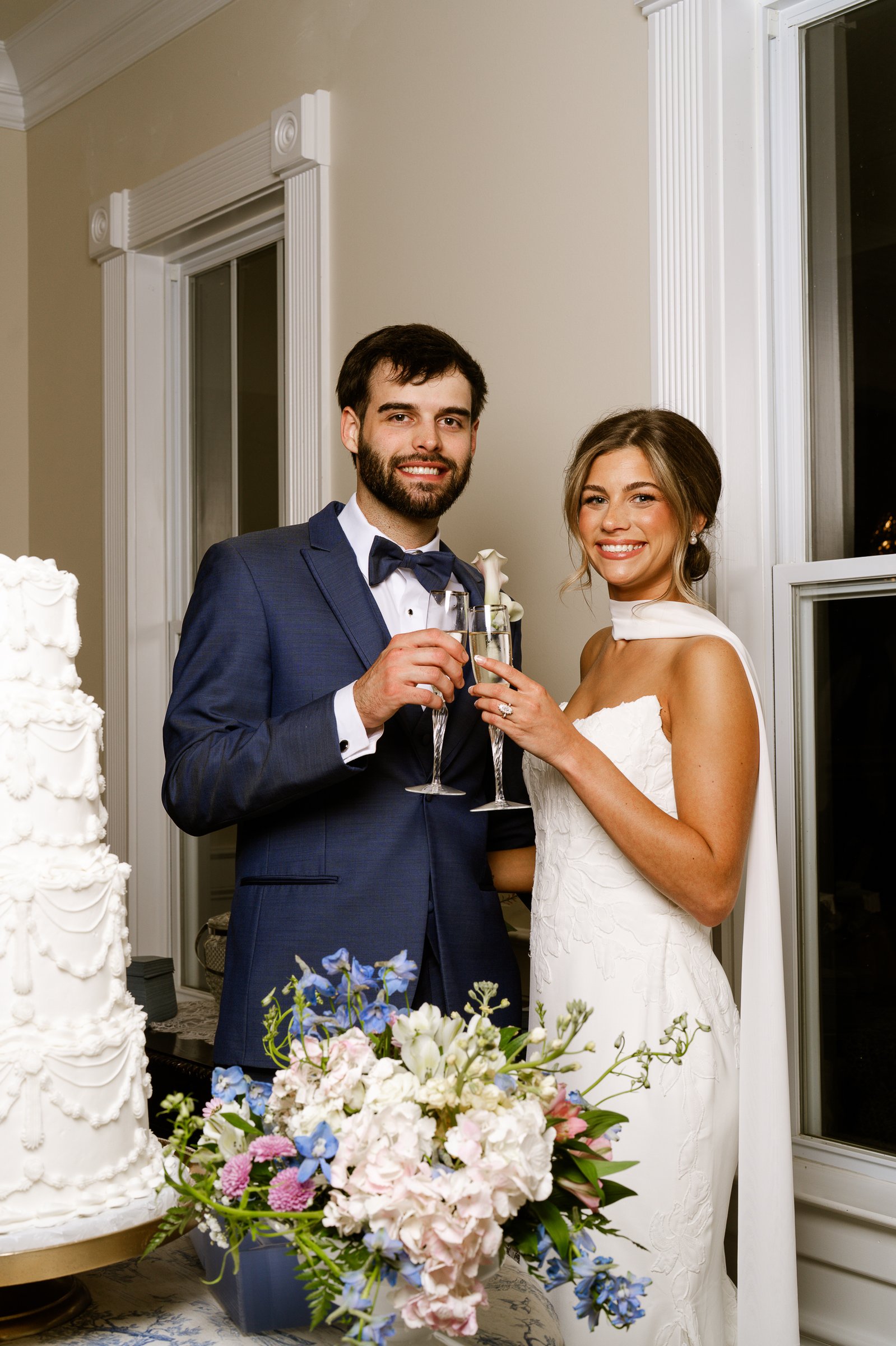 Matthew and Joley hold champagne flutes beside a tall white cake and a bouquet of pink and blue flowers.
