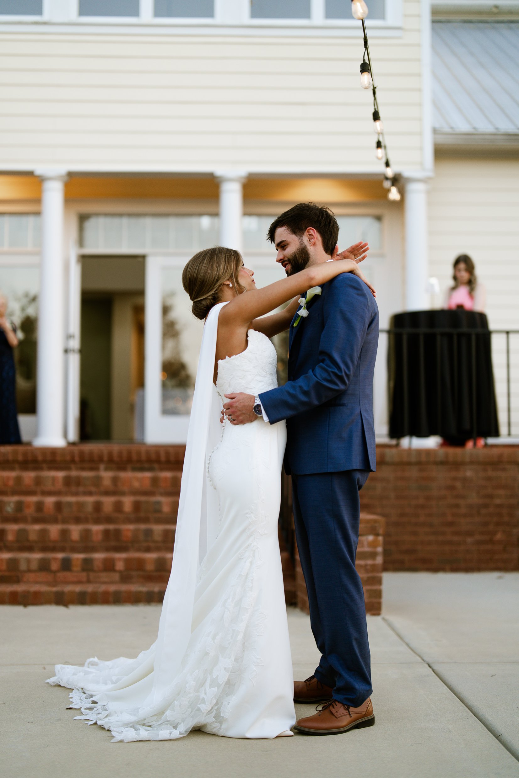 Joley and Matthew embrace on a patio in front of a house with string lights, her train spread behind her.