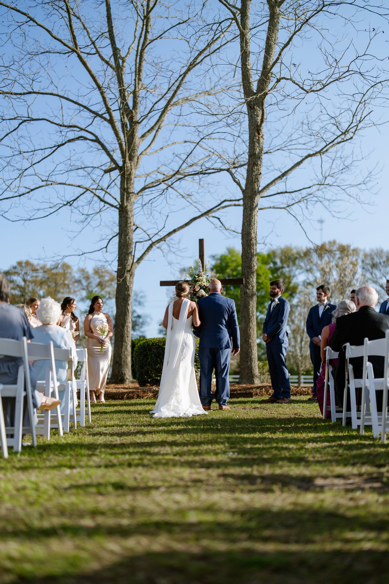 Seen from behind, Joley walks down a grass aisle between white chairs toward a wooden cross under bare trees.