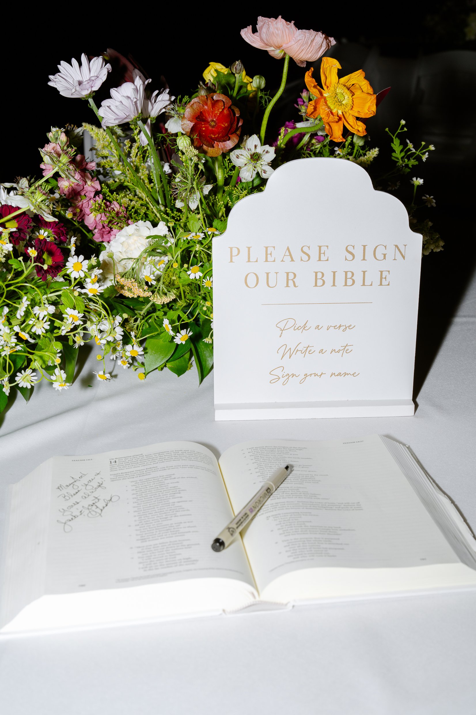 A Please sign our Bible sign beside an open Bible and a pen, with wildflowers behind.