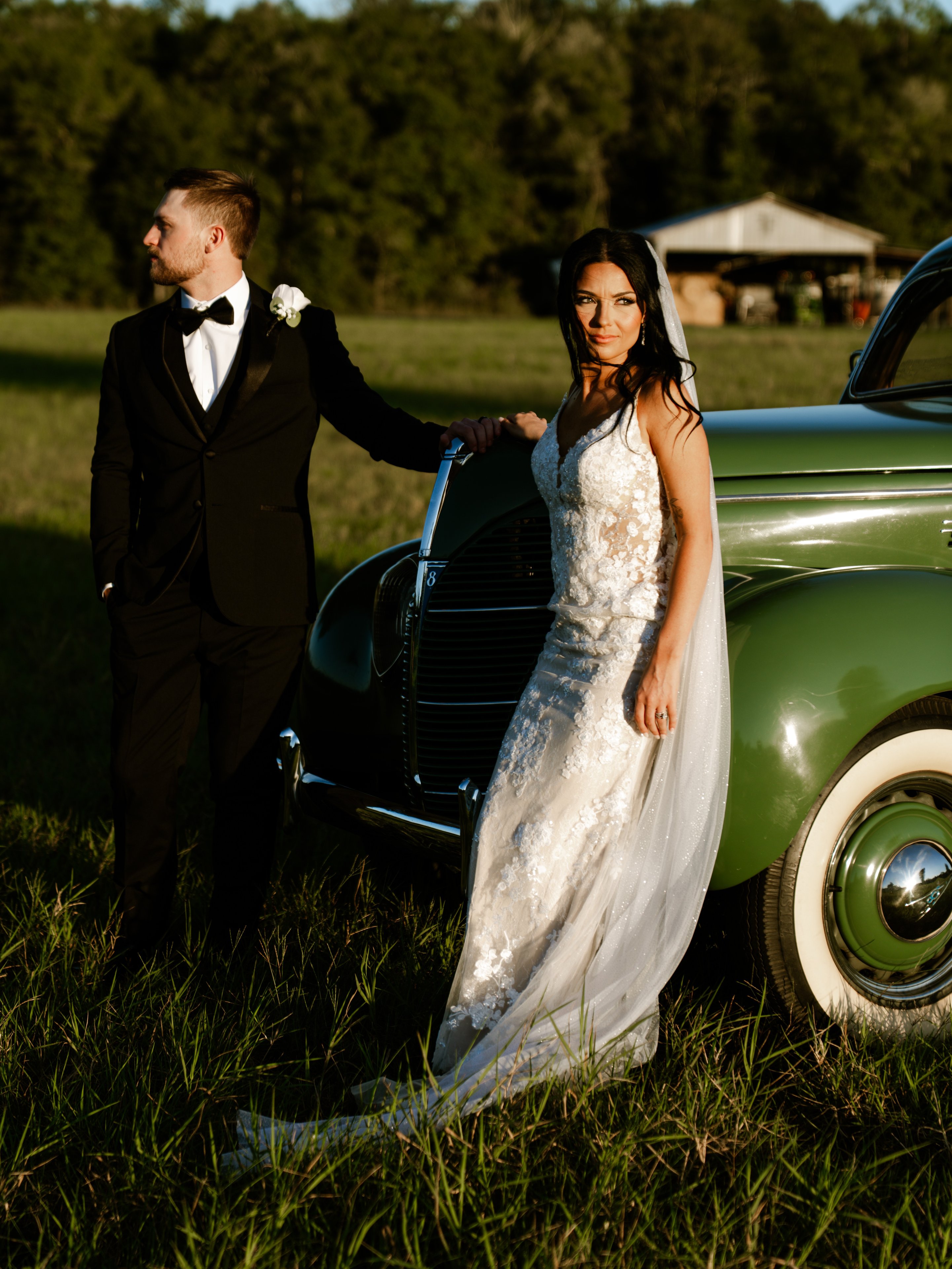Corey leaning on a green vintage car in a field while Kenadee stands next to it in her dress and veil.
