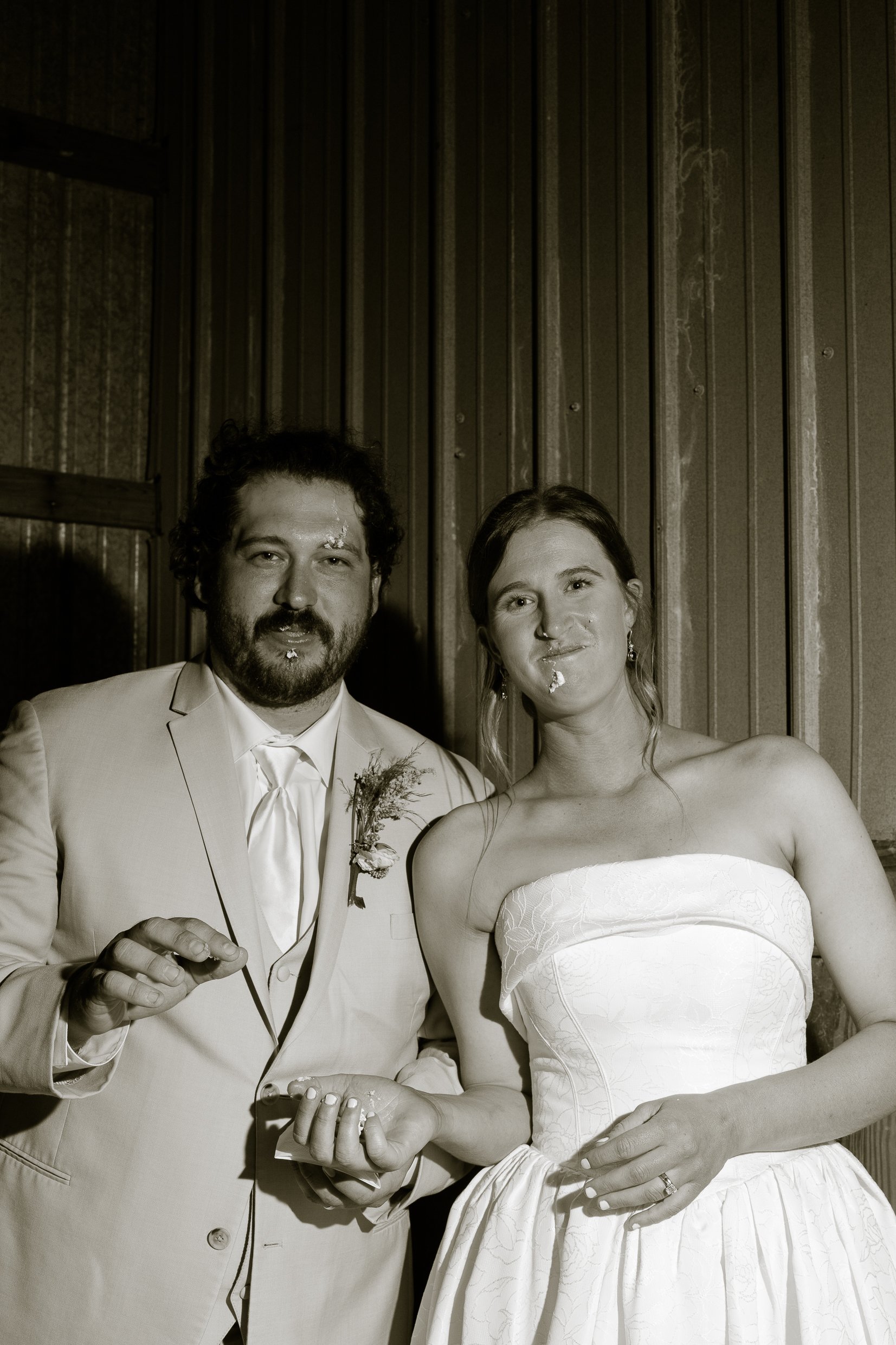 Elizabeth and Dustin look at the camera with cake on their faces, holding hands, in a warm toned black and white.