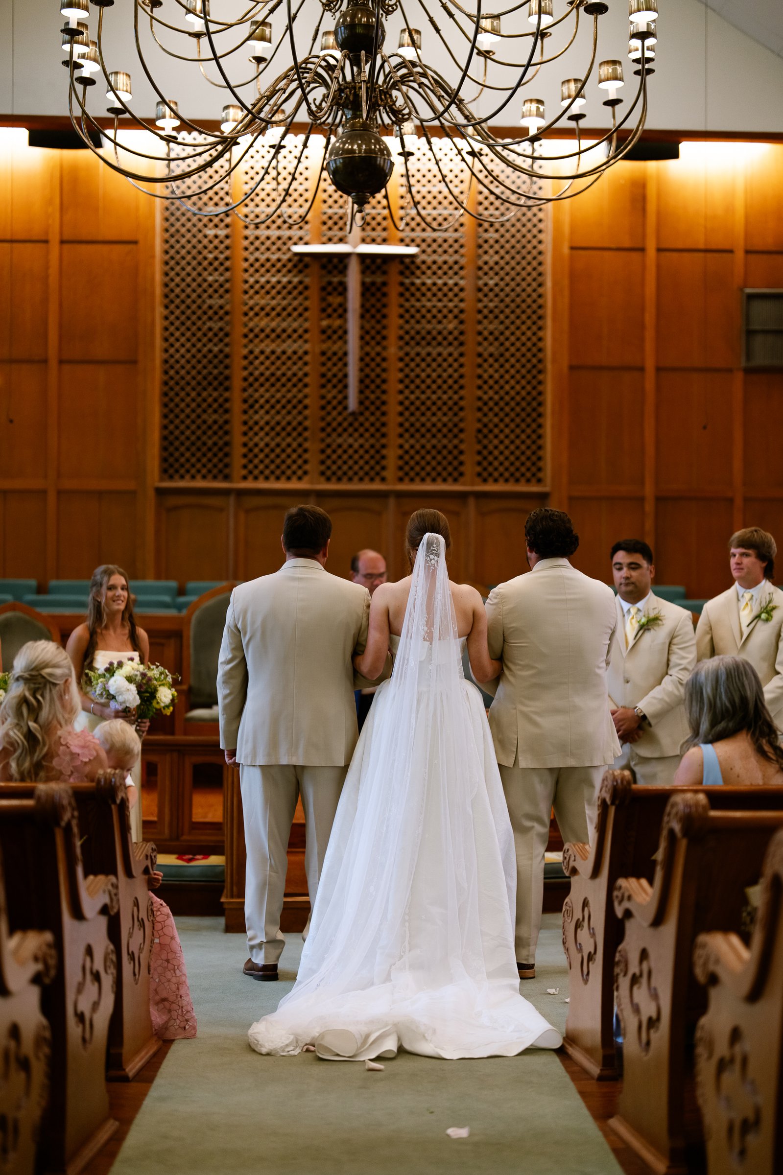 Elizabeth stands at the front of the church with her veil down her back, between two men in tan suits, with guests in the pews on both sides.