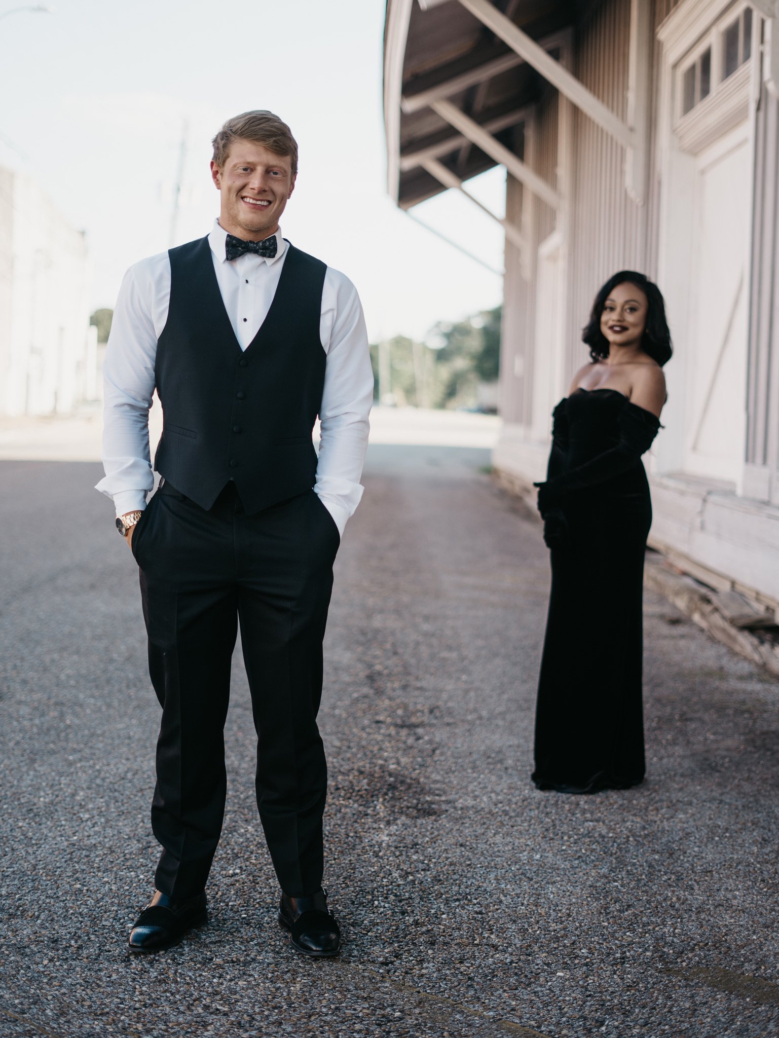 Jason stands with his hands in his pockets on a street beside a wooden building while Ashley stands farther back in a black velvet dress.