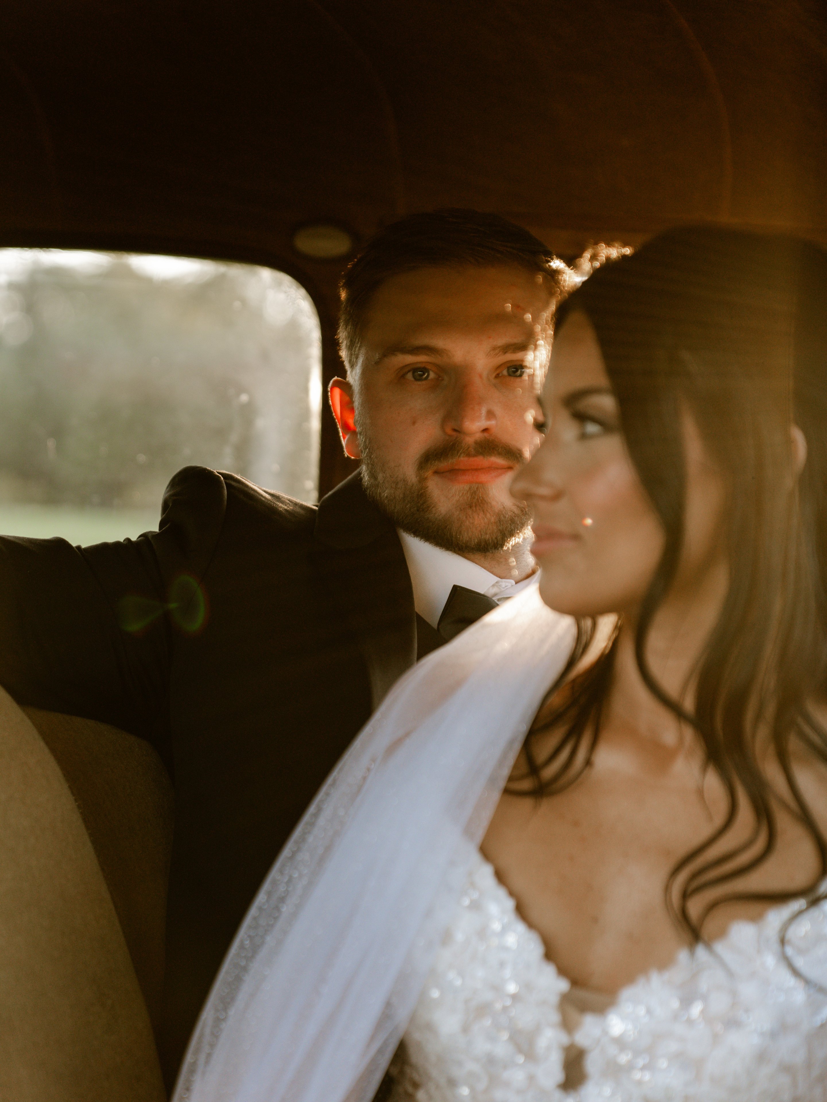 Kenadee and Corey close together in the back seat of a vintage car, her veil draped over her shoulder.