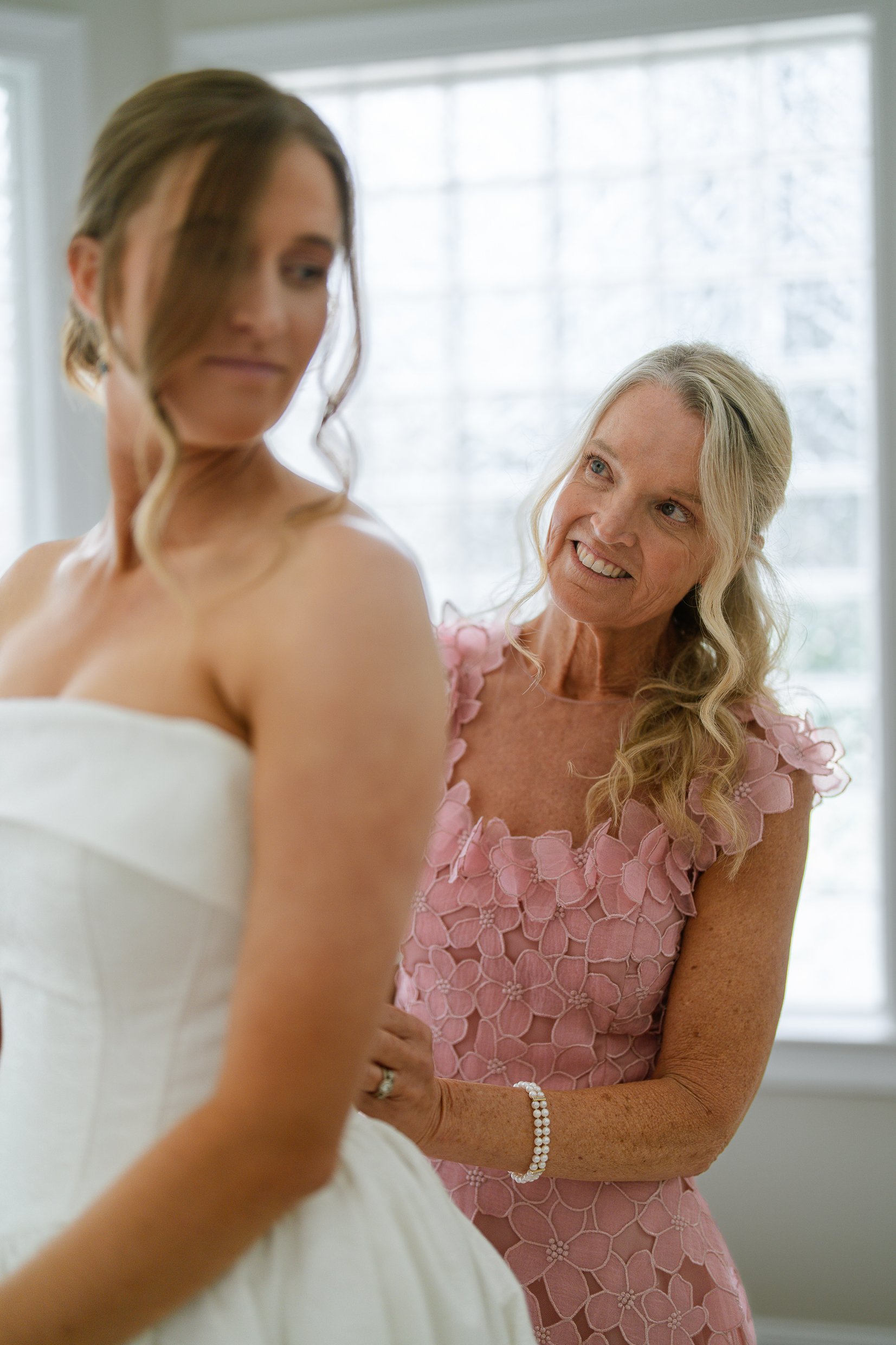 A woman in a pink lace dress smiles at Elizabeth as she fastens the back of her gown.
