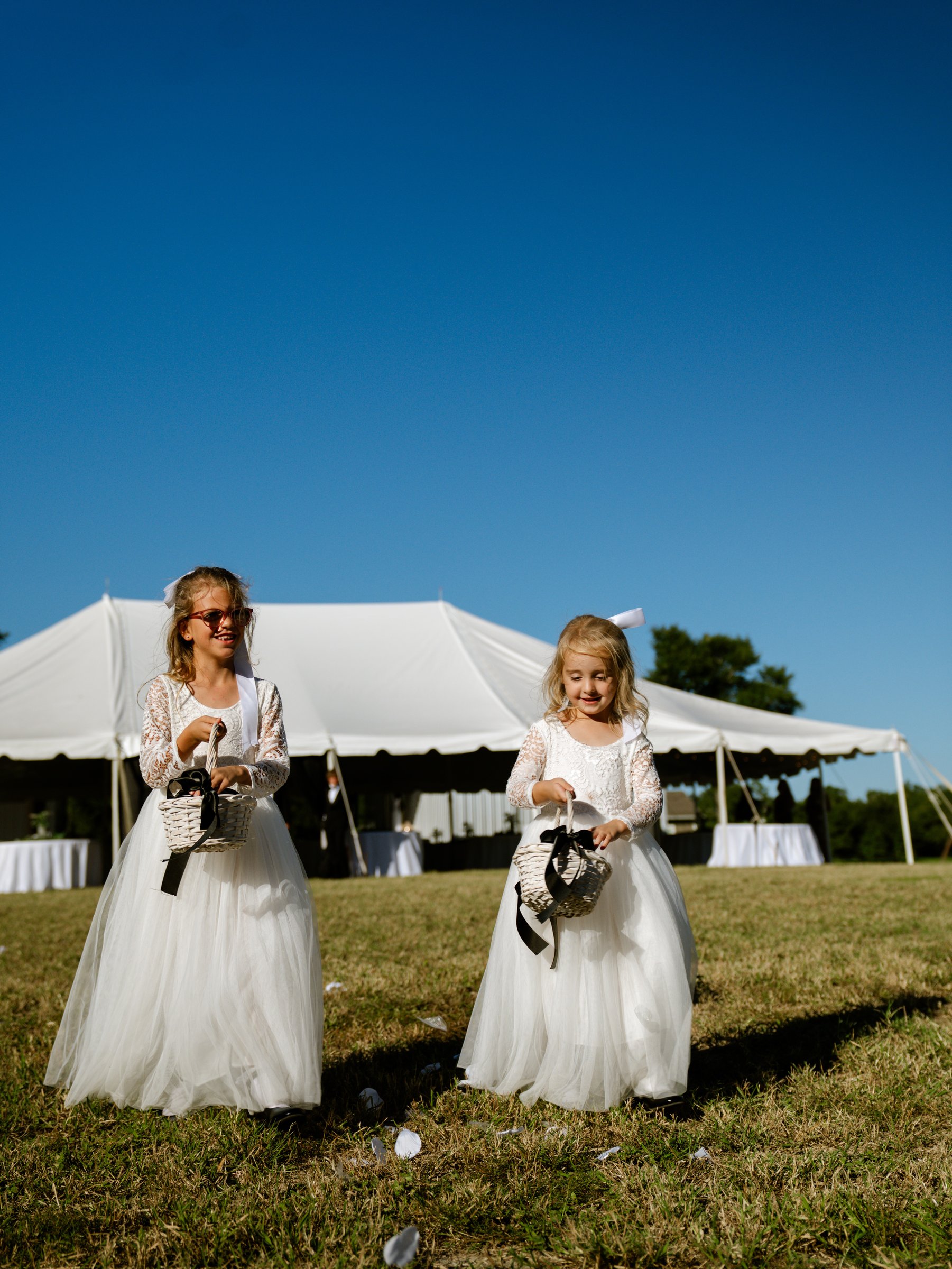 Two girls in white dresses holding small baskets stand on a lawn in front of a white tent.