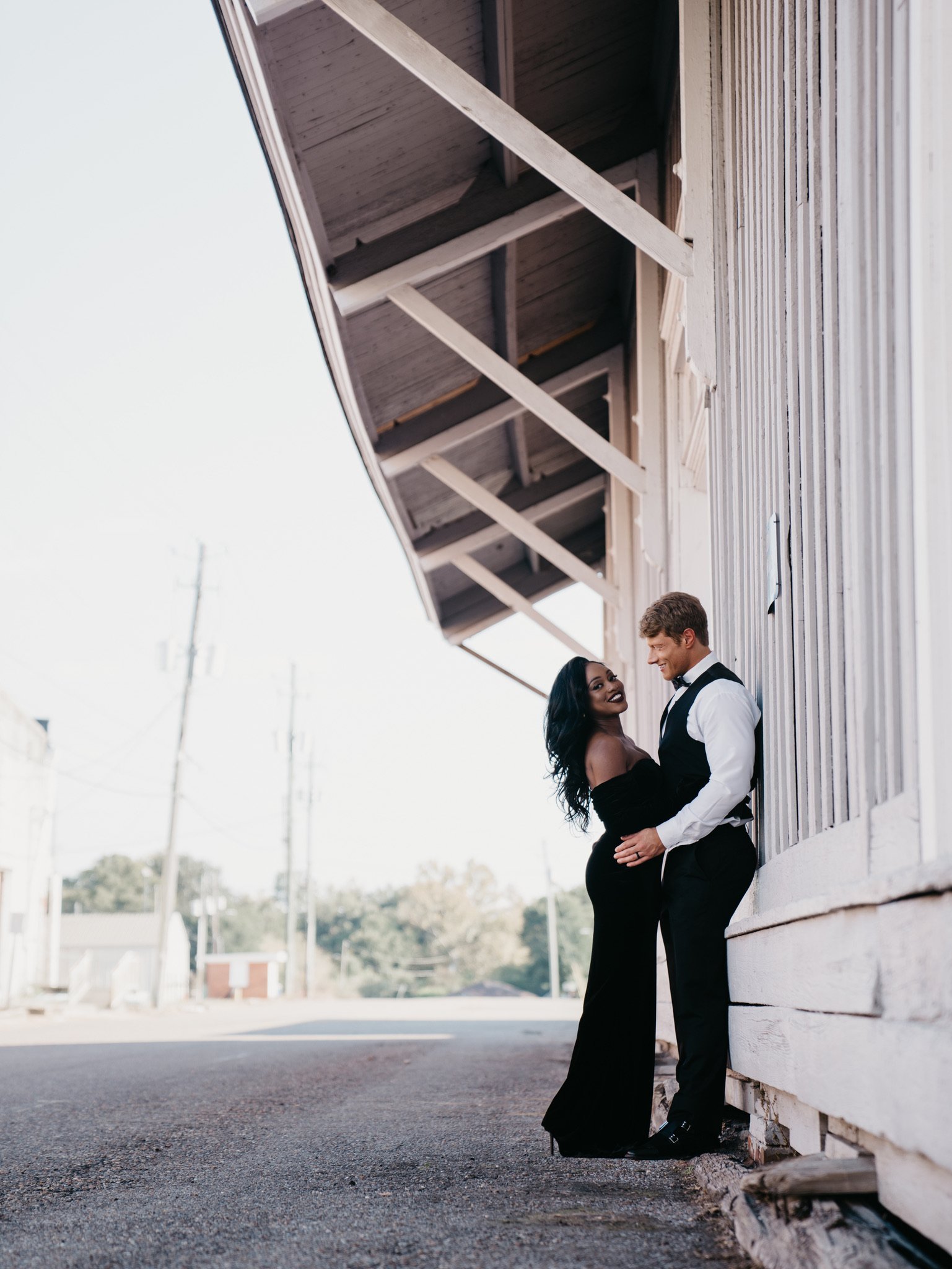 Ashley and Jason stand close against the white wall of an old wooden building, small in the frame on an empty street.