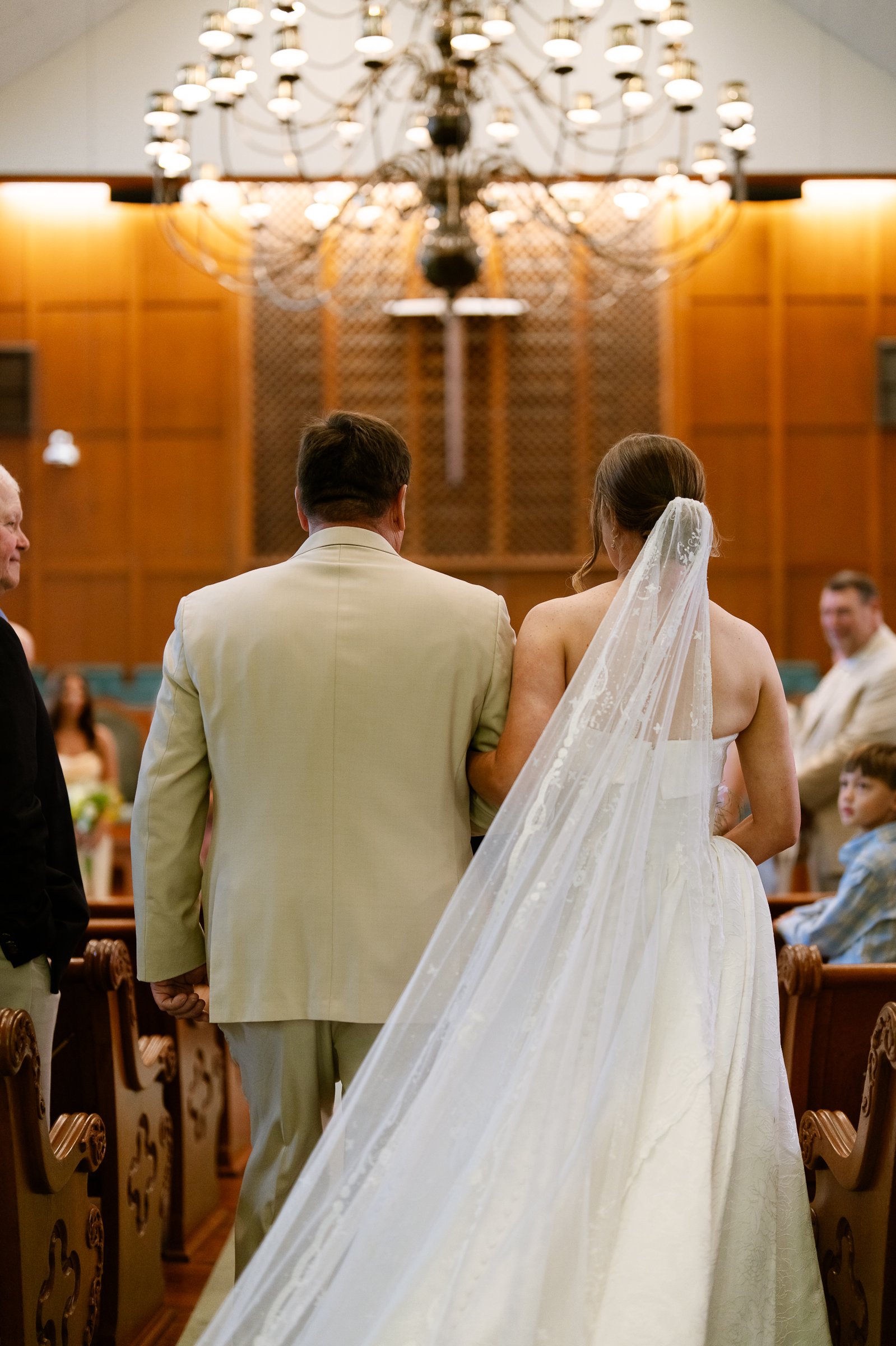 Elizabeth walks up the aisle on the arm of a man in a tan suit, her long veil trailing behind them under a chandelier.