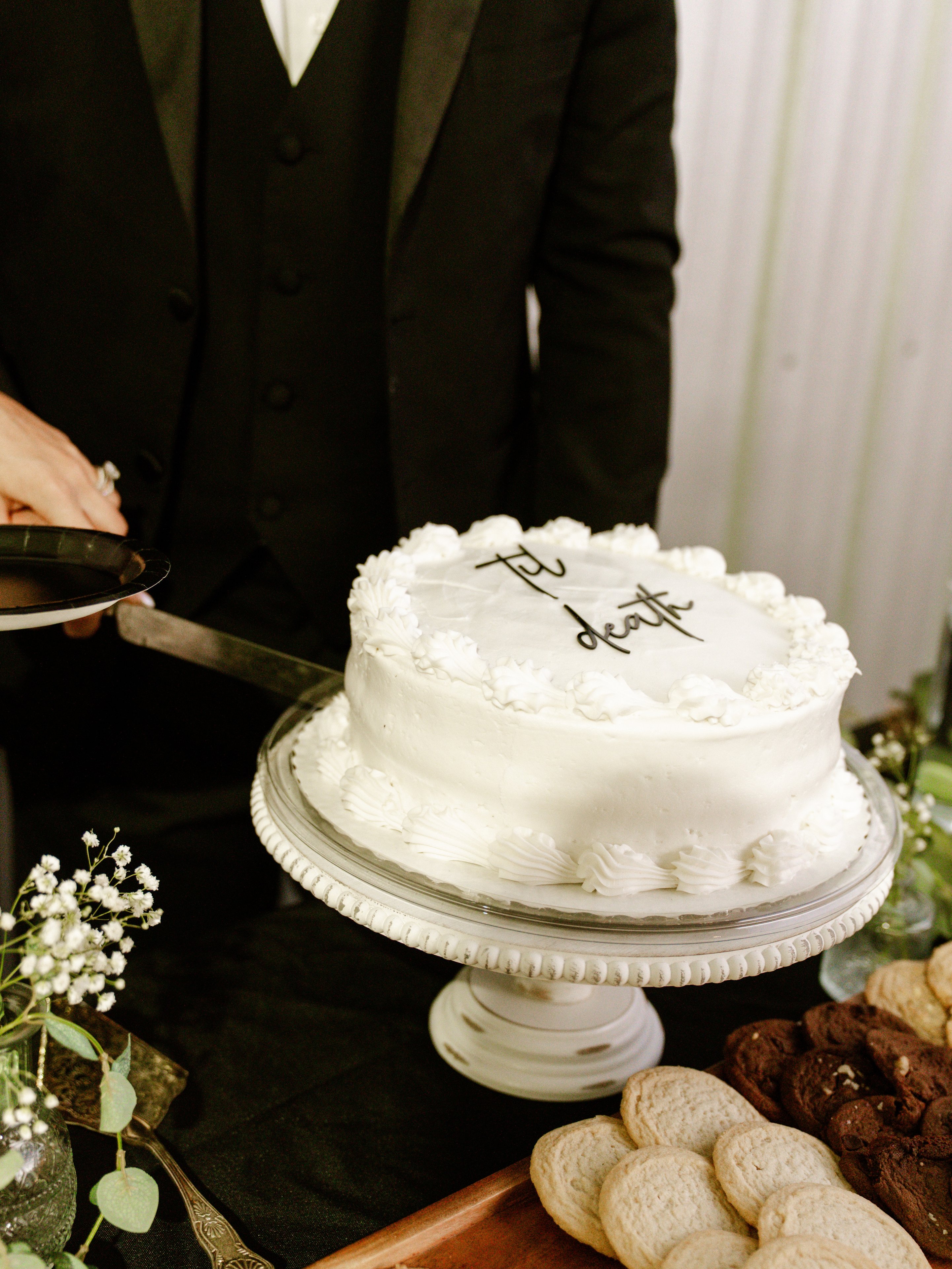 A white cake with writing on top on a stand, with a knife cutting into it and a plate of cookies beside it.