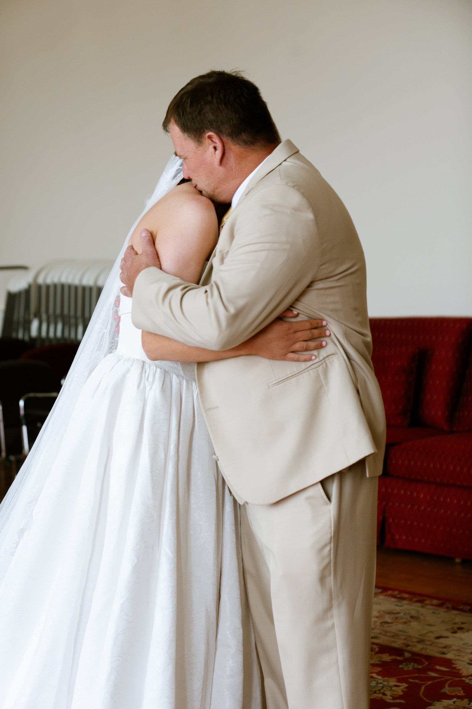 Elizabeth's father kisses the top of her head as they hold each other in a bright room with a red sofa behind them.