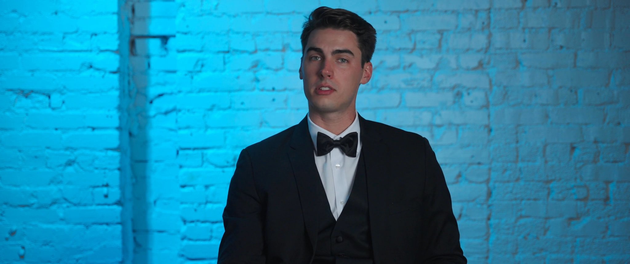 Jake in his tuxedo against a blue-lit wall. Still from their Midsummer Productions wedding film.
