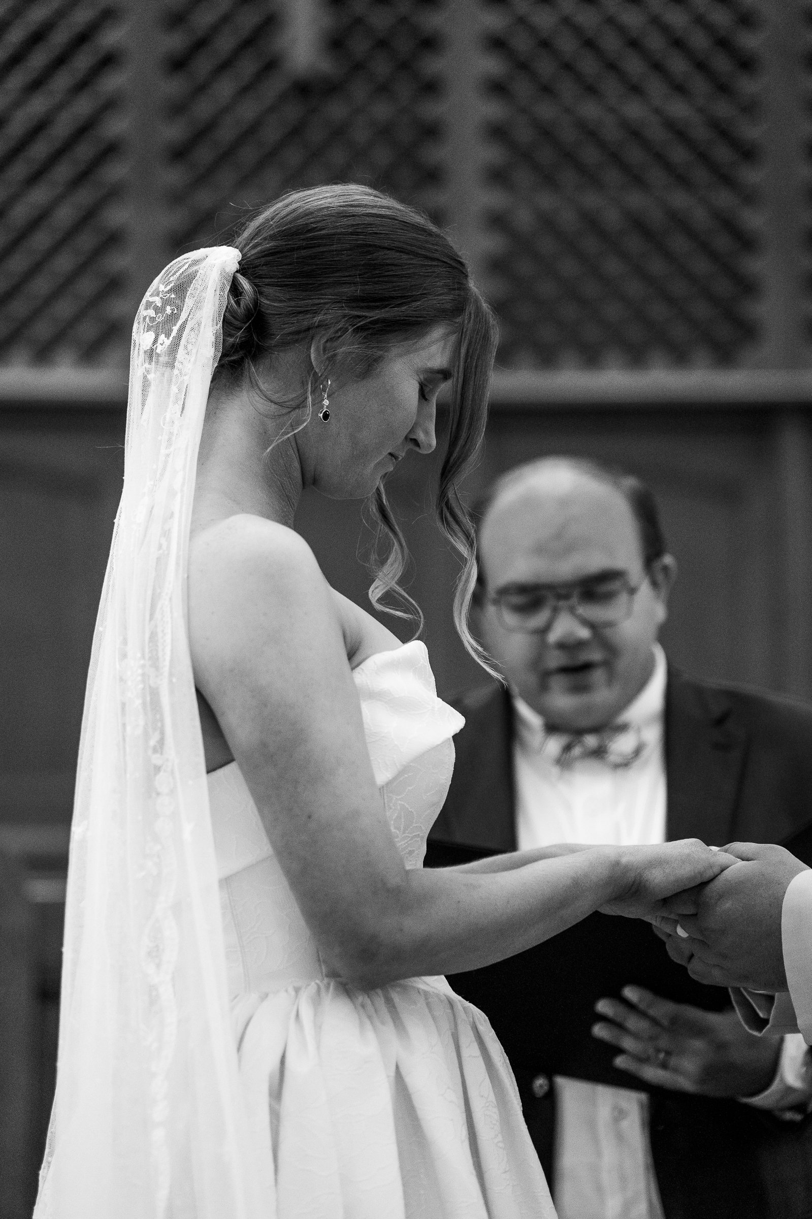 In black and white, Elizabeth looks down at their joined hands during the ceremony, her veil over her shoulder and the officiant behind her.