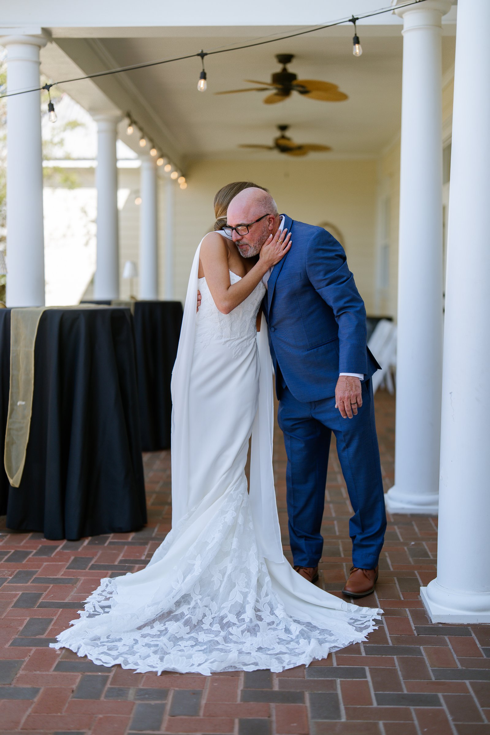 Joley hugs a man in a blue suit on a brick porch with white columns, her lace train spread behind her.