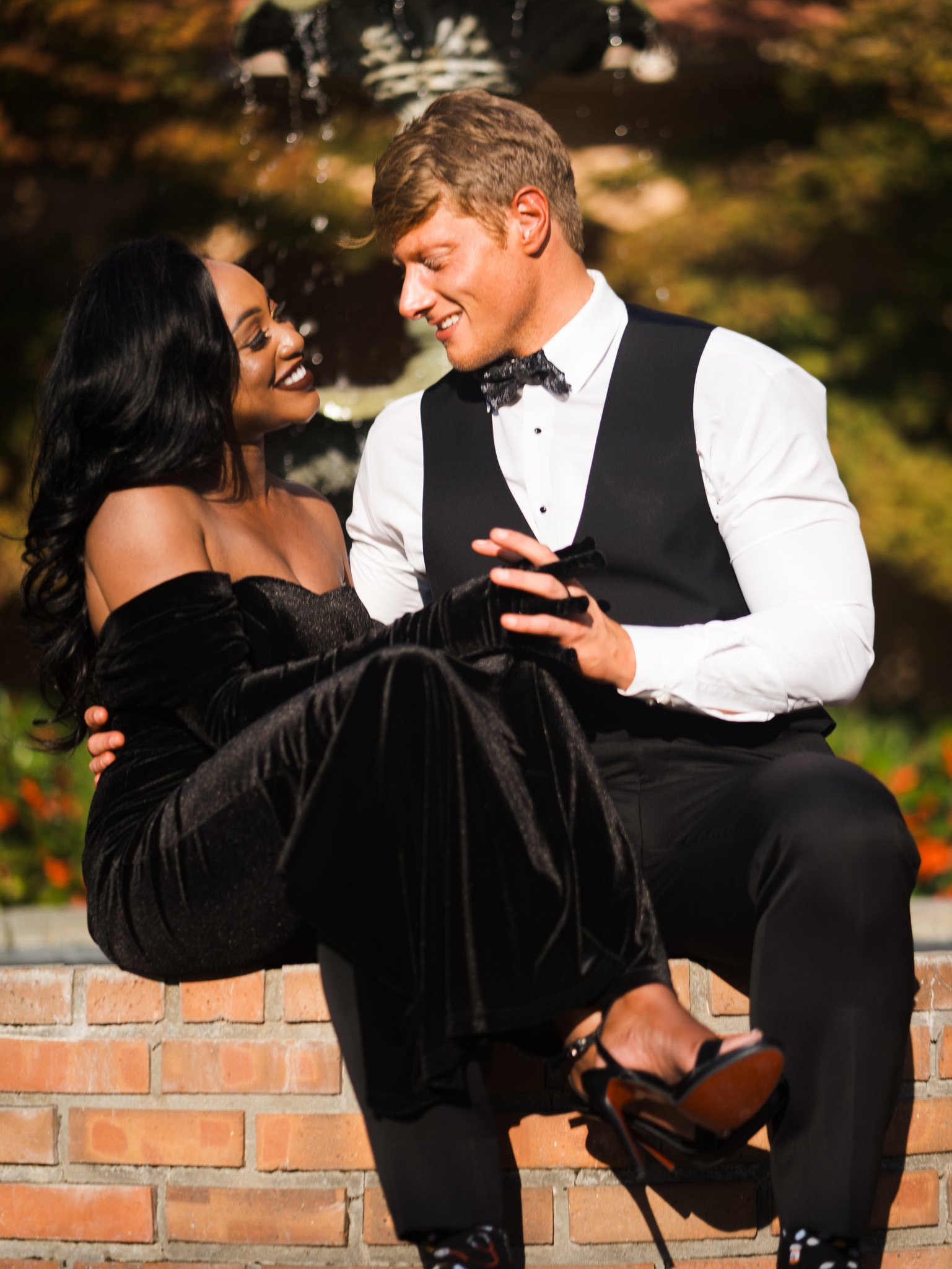 Ashley sits on Jason's lap on a low brick wall, both smiling, with trees and orange flowers behind them.