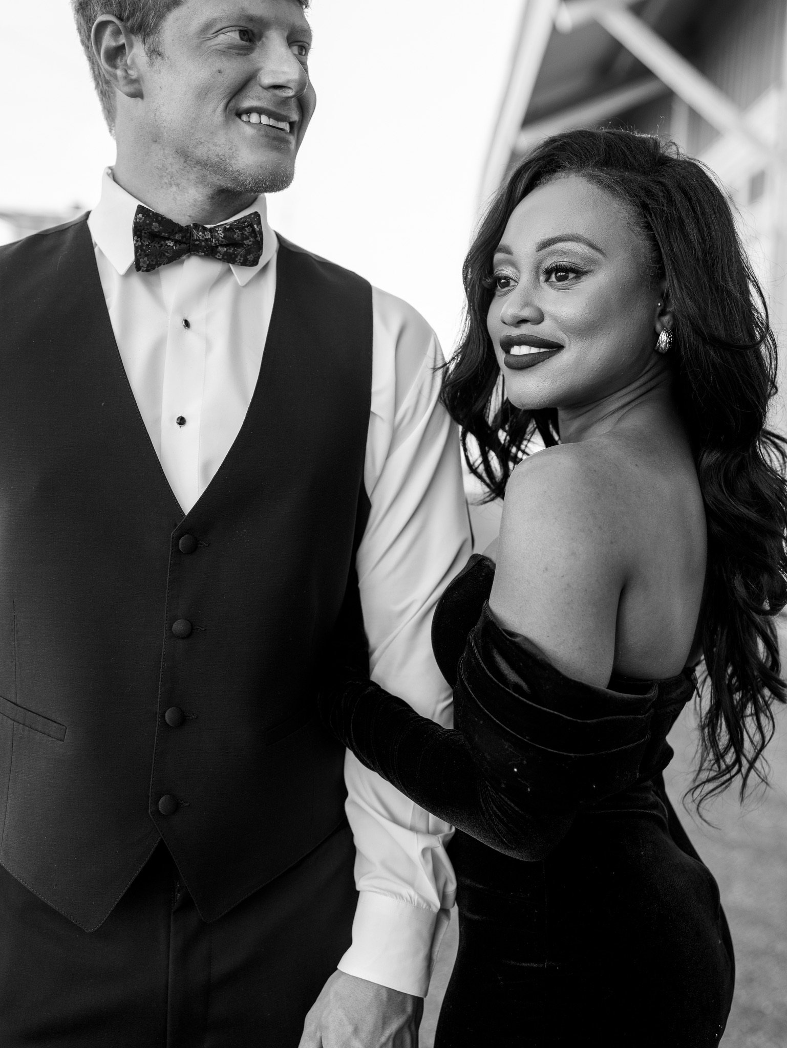 A black and white portrait of Jason in a vest and bow tie and Ashley looking back over her shoulder, both smiling.