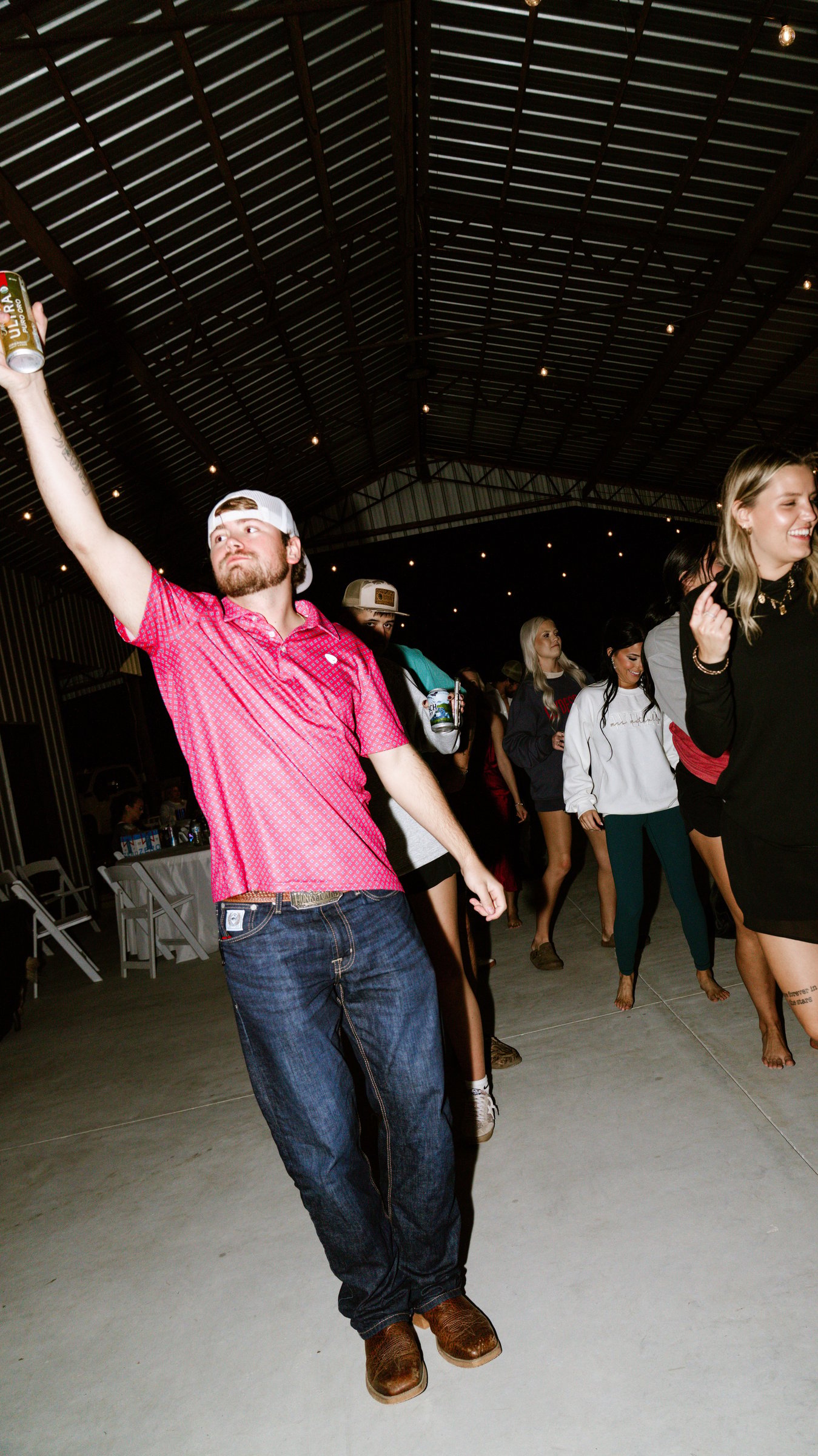 A man in a pink shirt with one arm raised high on the dance floor at night, with guests behind him.