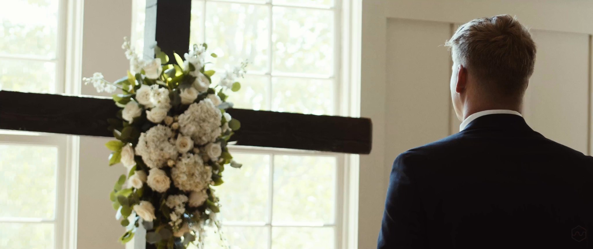 Caleb waits in front of the flower-covered cross for his first look with Madalyn. Still from their Midsummer Productions wedding film.