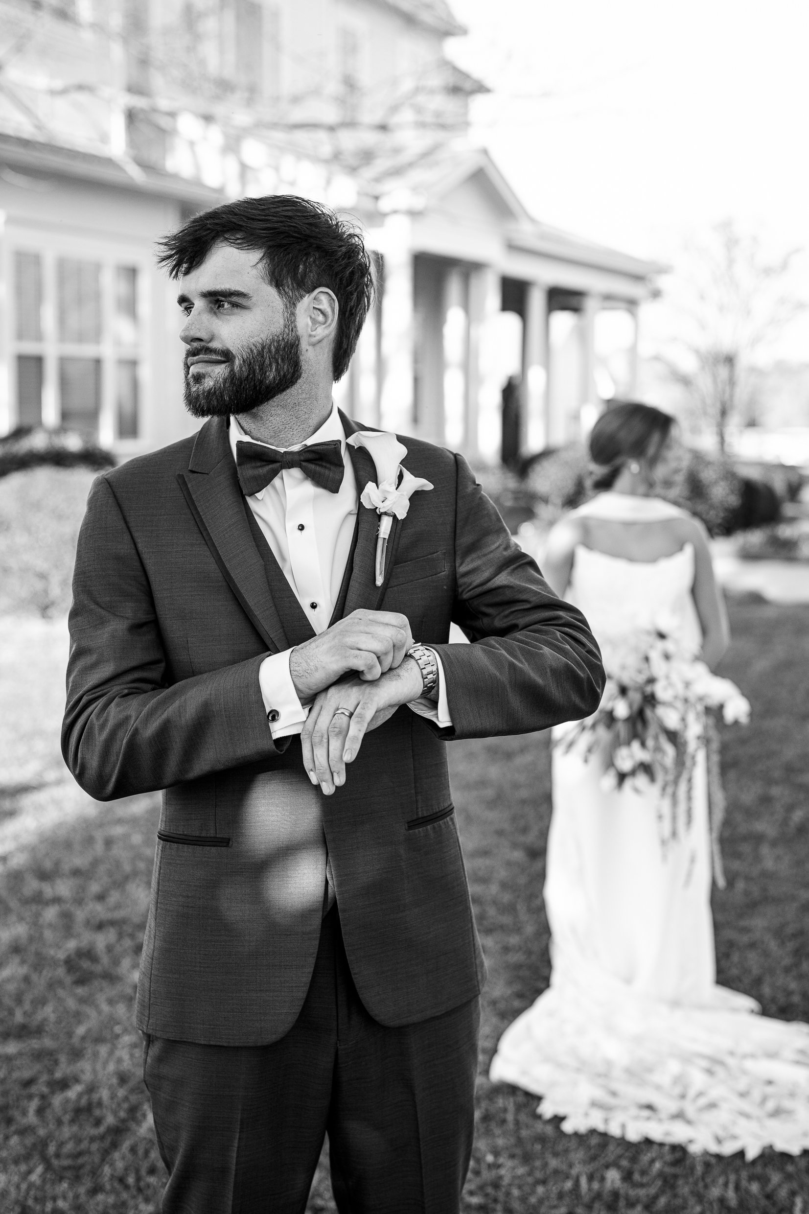 Black and white photo of Matthew in a bow tie checking his watch, with Joley in her dress blurred behind him.